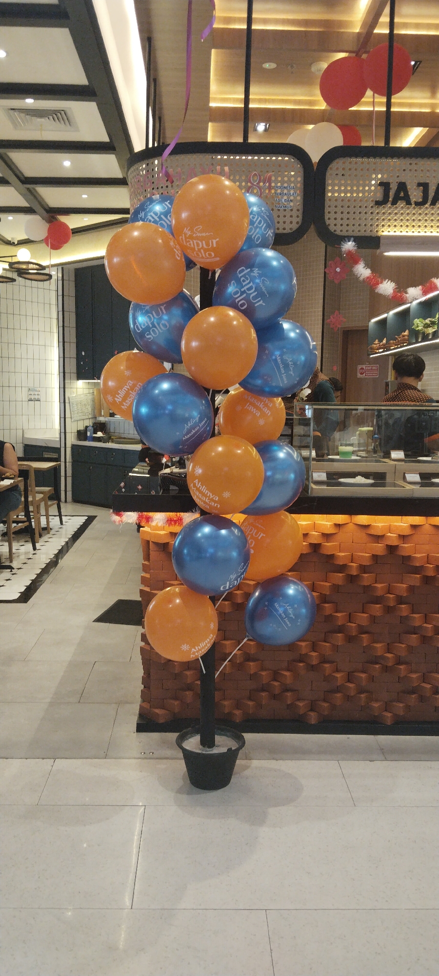 In Semarang, Indonesia, a tall display of orange and blue balloons stands in front of a brick-patterned counter. The balloons are arranged in a tiered fashion, with the majority featuring text that appears to be in Indonesian. Some of the visible phrases include "dapur solo," "Masakan Jawa," and "Ahlinya Masakan." The balloons are tethered to a black pole planted in a black pot filled with a gray substance, likely cement. Behind the balloons, the counter is illuminated by warm lighting, and a display case with food items is visible, suggesting this is a food establishment. People are seen in the background, including a server behind the counter and patrons seated at tables on the left side of the image. The flooring is a light-colored tile. A decorative red and white garland is draped across the top of the counter.