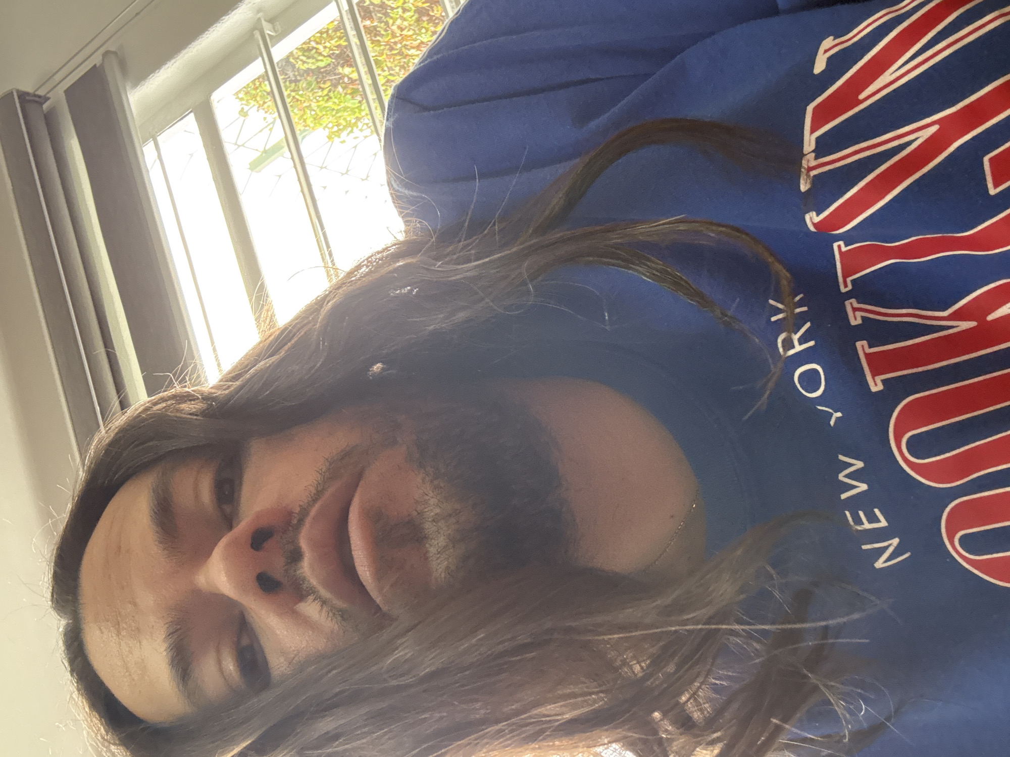 A close-up, rotated selfie captures an individual with long, dark hair and a beard, looking upwards towards the camera with a neutral to slightly smiling expression. The person wears a royal blue hooded sweatshirt featuring prominent red and white lettering. The visible text on the garment includes "NEW YORK" in white script, along with portions of larger red letters, likely part of a team or city name given the design and color scheme. A thin silver necklace is visible around the person's neck.
The background shows a bright window with white frames, through which green foliage and a light-colored fence or lattice are visible. Light-colored curtains are partially drawn on the left side of the window. The strong natural light coming through the window suggests the photo was taken indoors during the daytime. The image is set in Puebla, Mexico.