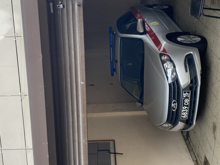 A silver Lada Granta sedan, identified as a Rosgvardiya (Russian National Guard) service vehicle, is parked indoors within what appears to be a garage or covered parking structure in Sochi, Russia. The vehicle features a red horizontal stripe along its side, a yellow-gold Rosgvardiya emblem on the front passenger door, and a blue emergency light bar mounted on its roof. The front license plate displays "6839 ОВ 15 RUS," indicating registration in the Republic of North Ossetia-Alania. An air freshener hangs from the rearview mirror.
The vehicle is situated beneath a partially open, horizontally slatted brown roll-up garage door. A padlock is visible on the right side of the door, securing it in its current elevated position. The interior walls of the structure are light-colored, possibly concrete or painted, with a rectangular window featuring a dark frame visible on the left wall; this window appears to be partially covered or obscured internally. The floor is covered with light-colored tiles or pavers. Above the roll-up door, a section of the building's exterior facade is visible, constructed with light grey square panels. A dark stool or similar object is partially visible in the lower right foreground, outside the garage opening. No individuals are present in the scene.