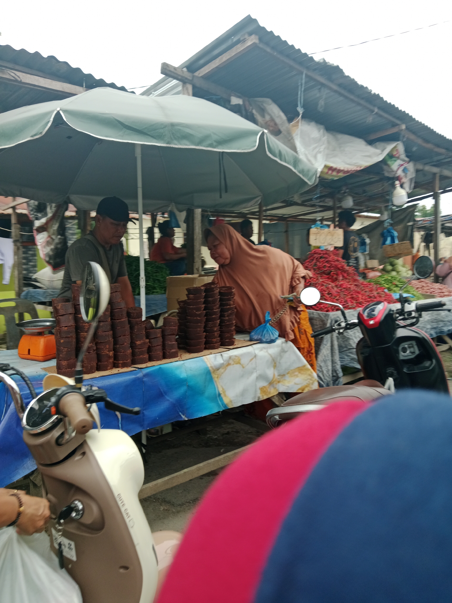 The image depicts a street market scene in the City of Binjai, Indonesia. In the foreground, a partially visible scooter with a brown seat and tan and white body is parked. A person's hand, adorned with a bracelet, is visible near the scooter's ignition. To the left of the scooter, a stall is set up under a large green umbrella. On the stall's table, which is covered with a blue and white patterned cloth, are stacks of dark brown palm sugar blocks. An orange weighing scale is also present. Behind the table, a man wearing a dark t-shirt and a black cap is visible, appearing to be attending the stall. To his right, a woman wearing a brown hijab and a peach-colored garment is also present. In the background, more market stalls are visible, with corrugated metal roofs. A large pile of red chilies is prominent, along with green vegetables and other produce. The overall environment suggests a bustling outdoor marketplace.