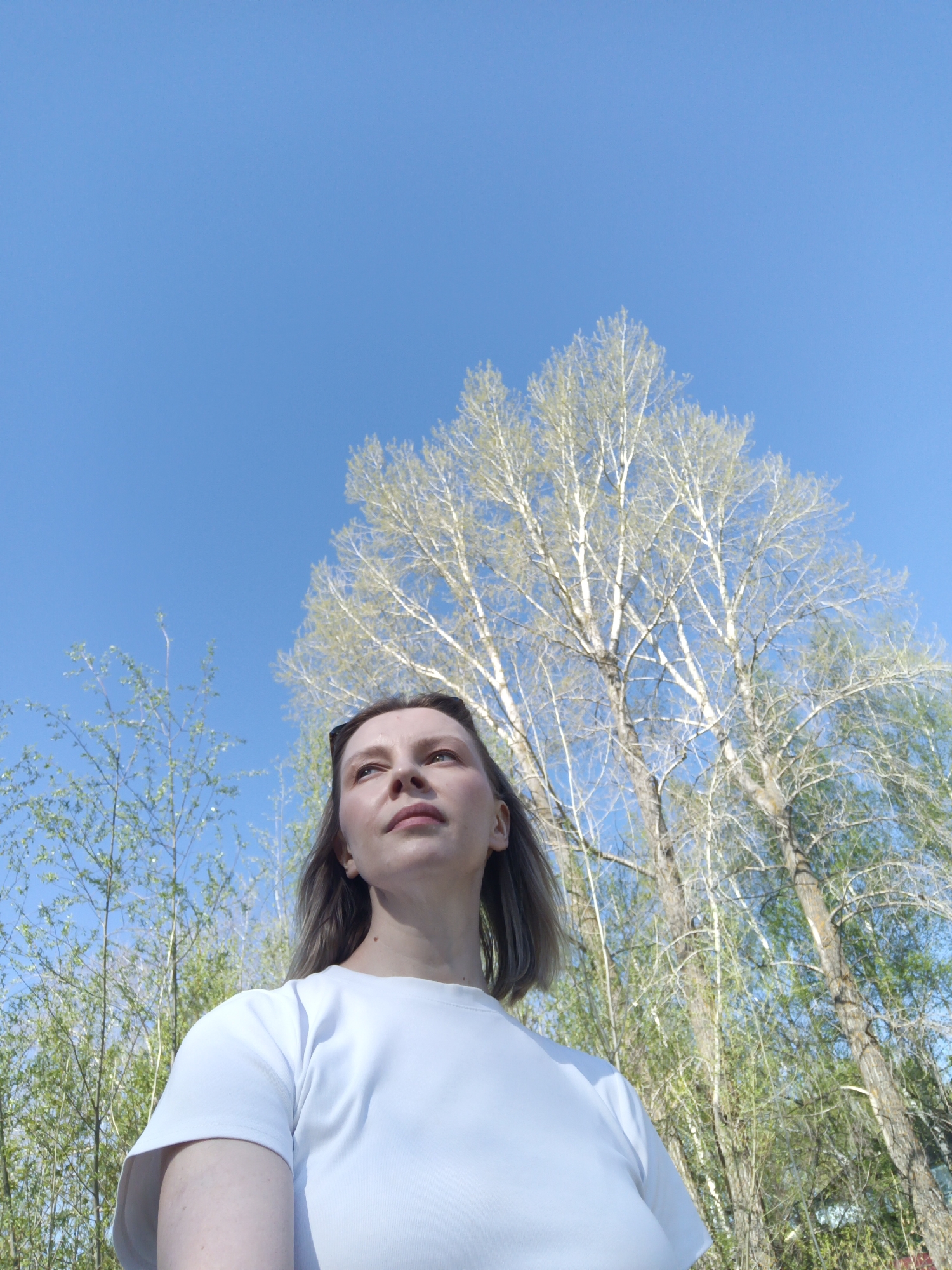 A low-angle shot features a woman outdoors against a bright blue sky with scattered bare trees and green foliage. She is dressed in a white T-shirt and looking upwards. The location is potentially Chaikovsky, Russia.