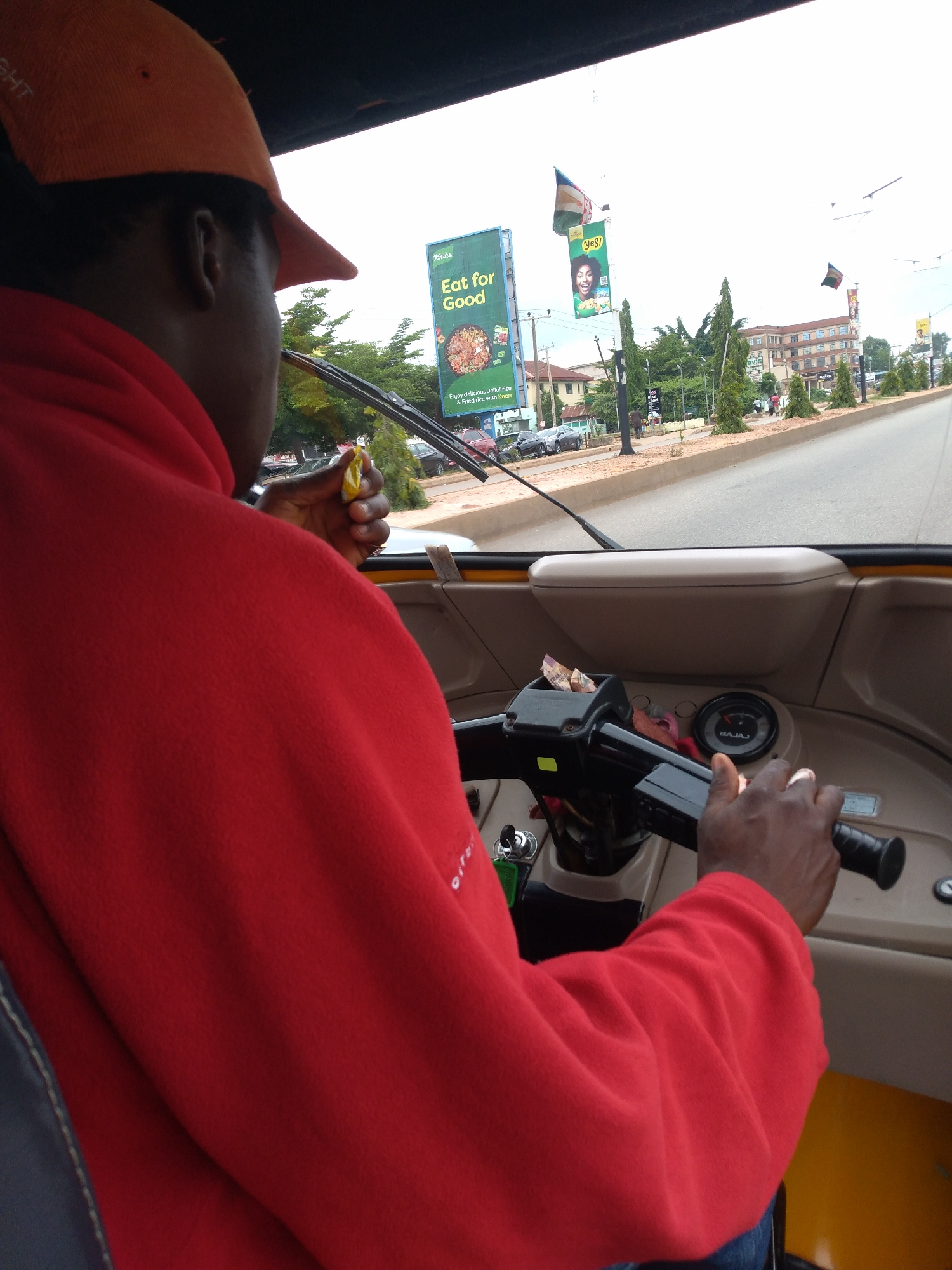 The image shows a first-person perspective from the driver's seat of a motorized three-wheeled vehicle, commonly known as a "keke" in Nigeria. The driver, wearing an orange cap and a red fleece jacket, is holding a small yellow object, possibly a piece of candy or medication, in their right hand. The vehicle's dashboard and steering mechanism are visible, including a speedometer labeled "BAJAJ". Through the windshield, a street scene in Jos, Nigeria, is depicted. Billboards line the roadside, one of which advertises "Eat for Good" with an image of food. Several cars and other vehicles are present on the road, and buildings and trees are visible in the background. The sky is overcast.