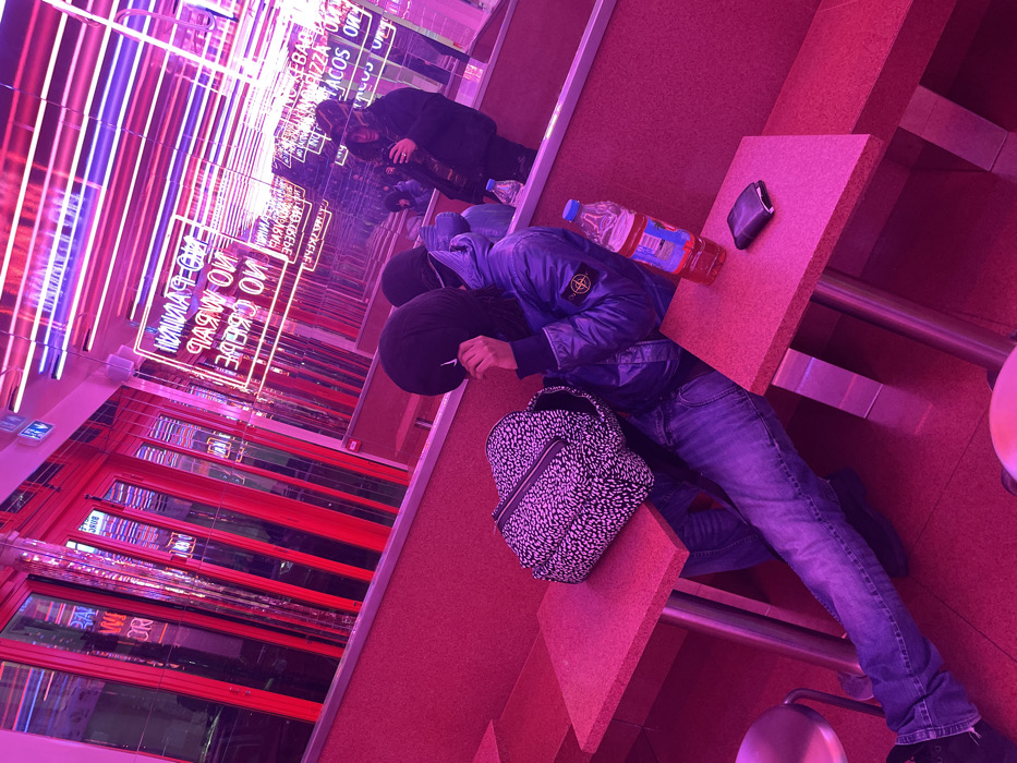 A person in a blue jacket and jeans sits at a pink table with a Dalmatian-print backpack and a water bottle. A black wallet rests on the table. Reflections in a mirrored wall show neon signs, including "NO CREPE, NO WAFFLE, NO PANINI", and windows with red accents. The scene is bathed in vibrant pink and purple hues.