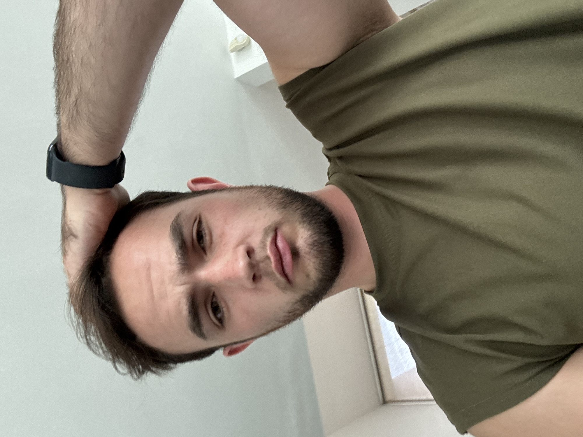 A man with dark hair and a beard is positioned in the center of the frame, looking down at the camera. He is wearing a dark green t-shirt and a black smartwatch on his left wrist. His left hand is placed on the back of his head. The background is primarily a plain, light-colored wall. There is a portion of a white window frame visible near his right shoulder. The location appears to be an indoor setting in San Bernardino Tlaxcalancingo, Mexico.