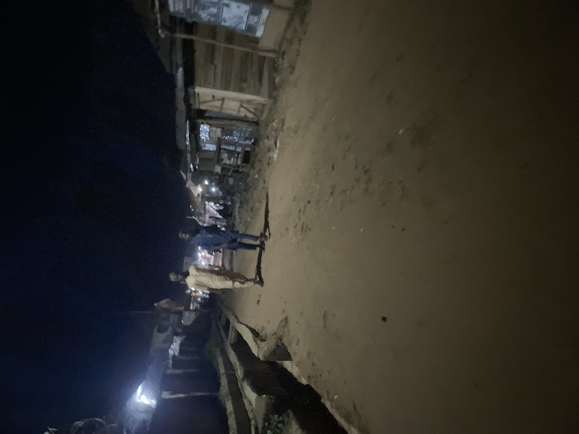 The media file shows a dimly lit, outdoor scene, likely in Lokoja, Nigeria, at night. The perspective is tilted, giving a sense of movement or instability. In the foreground, a muddy path or dirt road is visible, with a low concrete barrier running alongside it. Beyond the barrier, there's a dark, open space, possibly a body of water or a deep ditch.
Several figures are present in the scene, though their details are obscured by the low light. In the middle ground, two individuals appear to be walking or standing on the road. One person is wearing a light-colored garment, possibly a traditional outfit, and the other is in darker clothing. Further back, a group of people are gathered, some appearing to be interacting around what might be stalls or structures. Lights from these stalls are faintly visible, suggesting a market or gathering place. The overall atmosphere is dim and somewhat mysterious, typical of a night scene in an area with limited artificial lighting. The weather appears to be clear, as there are no indications of rain or fog. No visible text is present in the image.