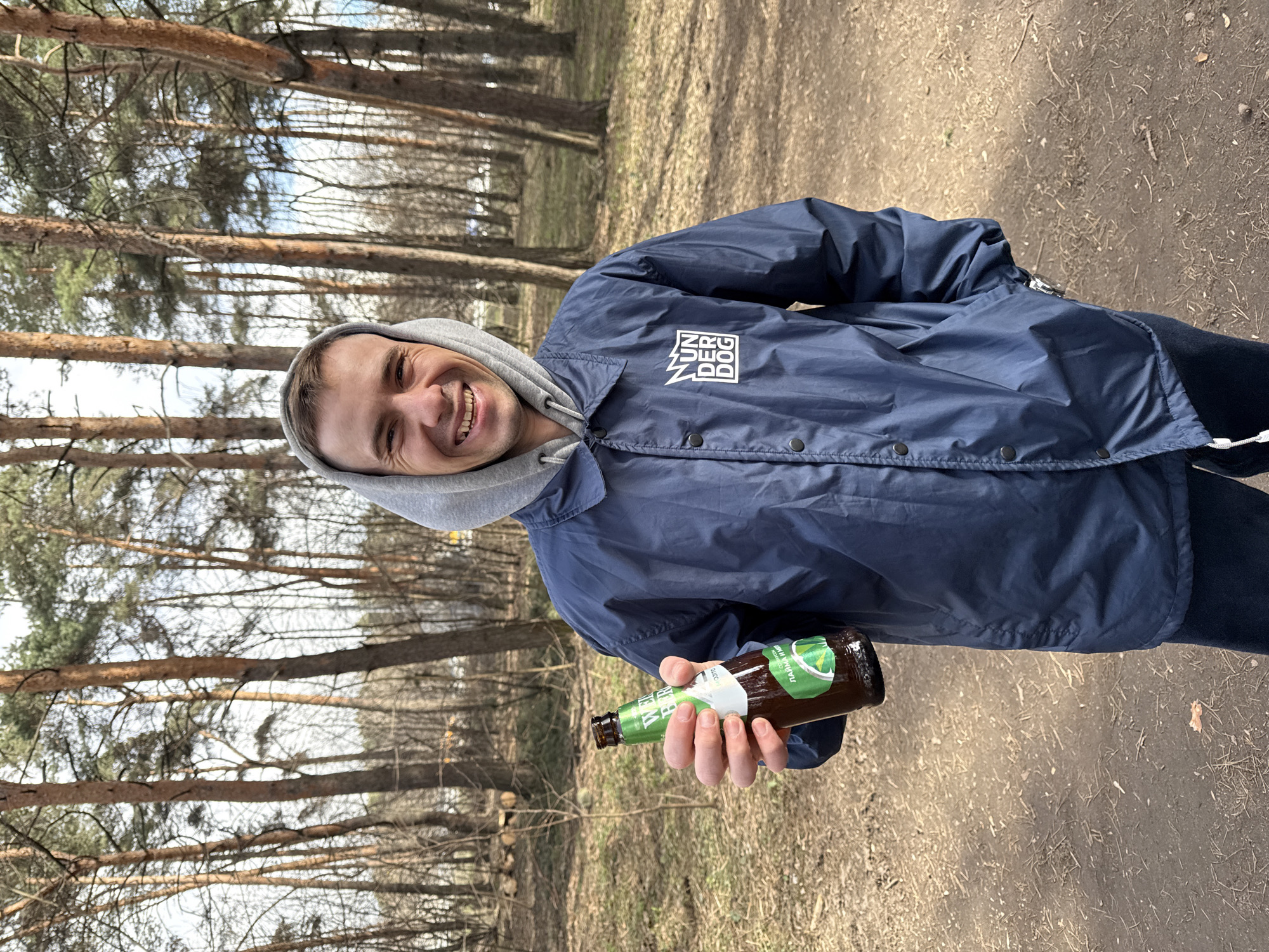 A man in a navy blue jacket with a gray hoodie stands in a park-like area, holding a brown beer bottle with a green label. The jacket has a logo on the left chest. He is smiling and looking directly at the camera. The background consists of tall, bare trees and a dirt path. The scene is illuminated by natural daylight. The beer bottle label has writing in Cyrillic and is likely a domestic brand. Based on the bare trees and the casual attire of the person, the weather is likely cool. Given the Cyrillic writing on the beer bottle label, the location may be the city of Moscow, Russia.