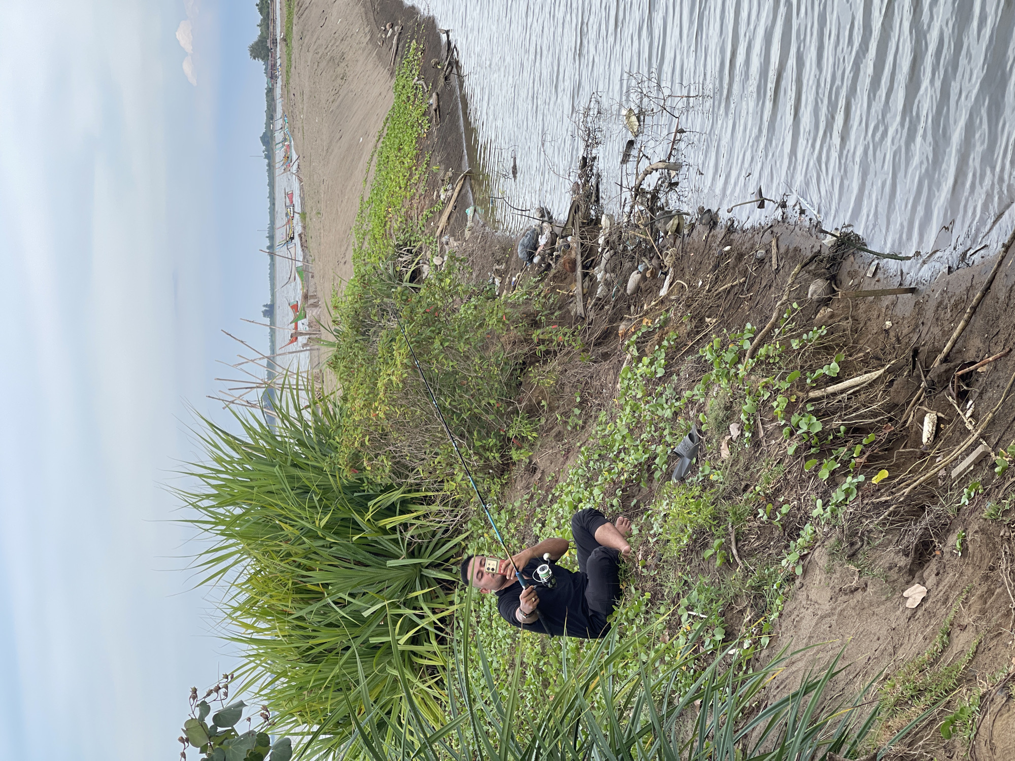A person is sitting on a grassy, sandy bank, fishing in a body of water. The person is wearing a black shirt and pants and is holding a fishing rod and a smartphone. The bank is covered in green vegetation and debris. In the background, there are several boats on the water and a shoreline with more vegetation. The sky is cloudy. This scene appears to be the Krueng Mane river, near Lhokseumawe, Indonesia.