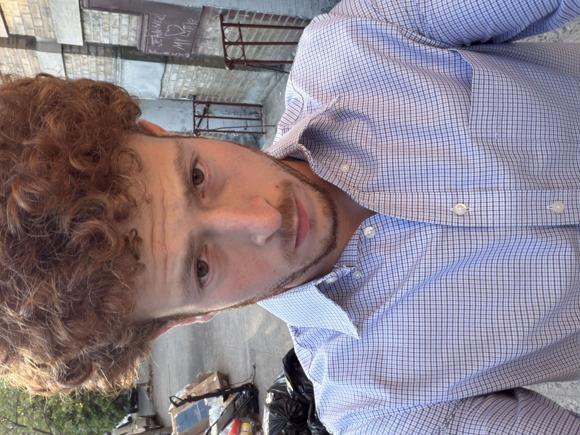 A person is facing the camera while standing outdoors. The person is wearing a button-down shirt with a checked pattern of light blue and dark blue lines. They have light skin, brown eyes, and curly red hair. A light beard is visible on their face. The person is standing on concrete. In the background, there are black trash bags, boxes, and greenery visible at the bottom of the image. Behind the person, a brick wall and metal fence is visible. A dark sign with light-colored writing can be seen behind the fence. This is likely New York City.