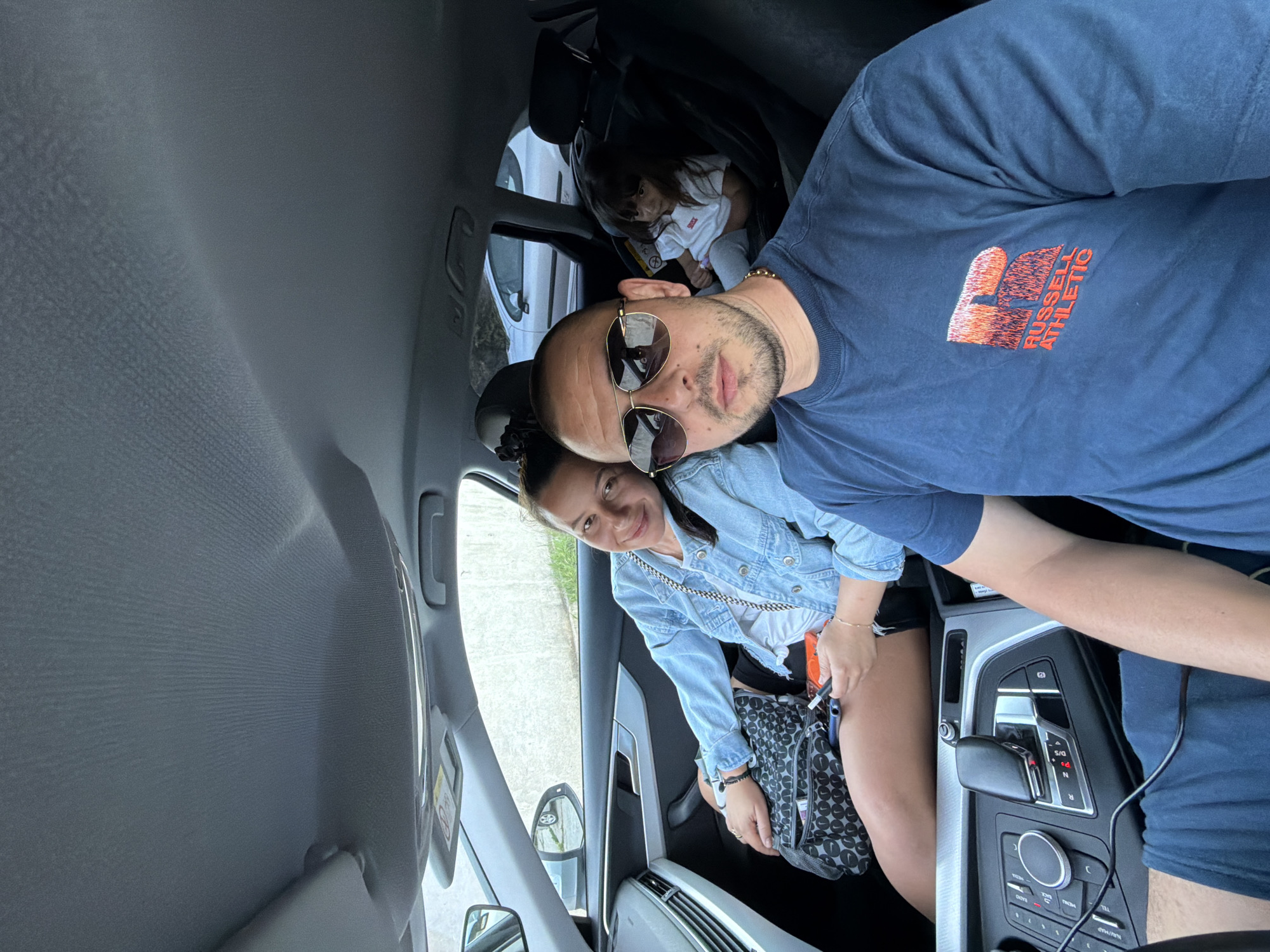 A family is taking a selfie in a car. A man wearing sunglasses and a blue t-shirt with the "Russell Athletic" logo on it is in the foreground. To his left, a woman in a denim jacket is smiling. In the back seat, a young child is seated in a car seat. The interior of the car, including the dashboard and gear shift, is visible. The family appears to be on the road, possibly traveling. The image does not contain any specific landmarks or indicators of the city of Burgas, Bulgaria.