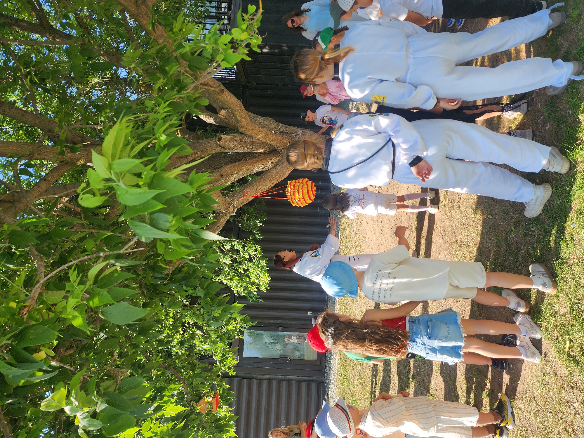 A group of children and adults are gathered outside, near a tree with a hanging orange and red striped lantern. Some of the adults and children are wearing white martial arts uniforms. The location appears to be near a corrugated metal building. Without further details, I cannot confirm that this is in Kazan, Russia.