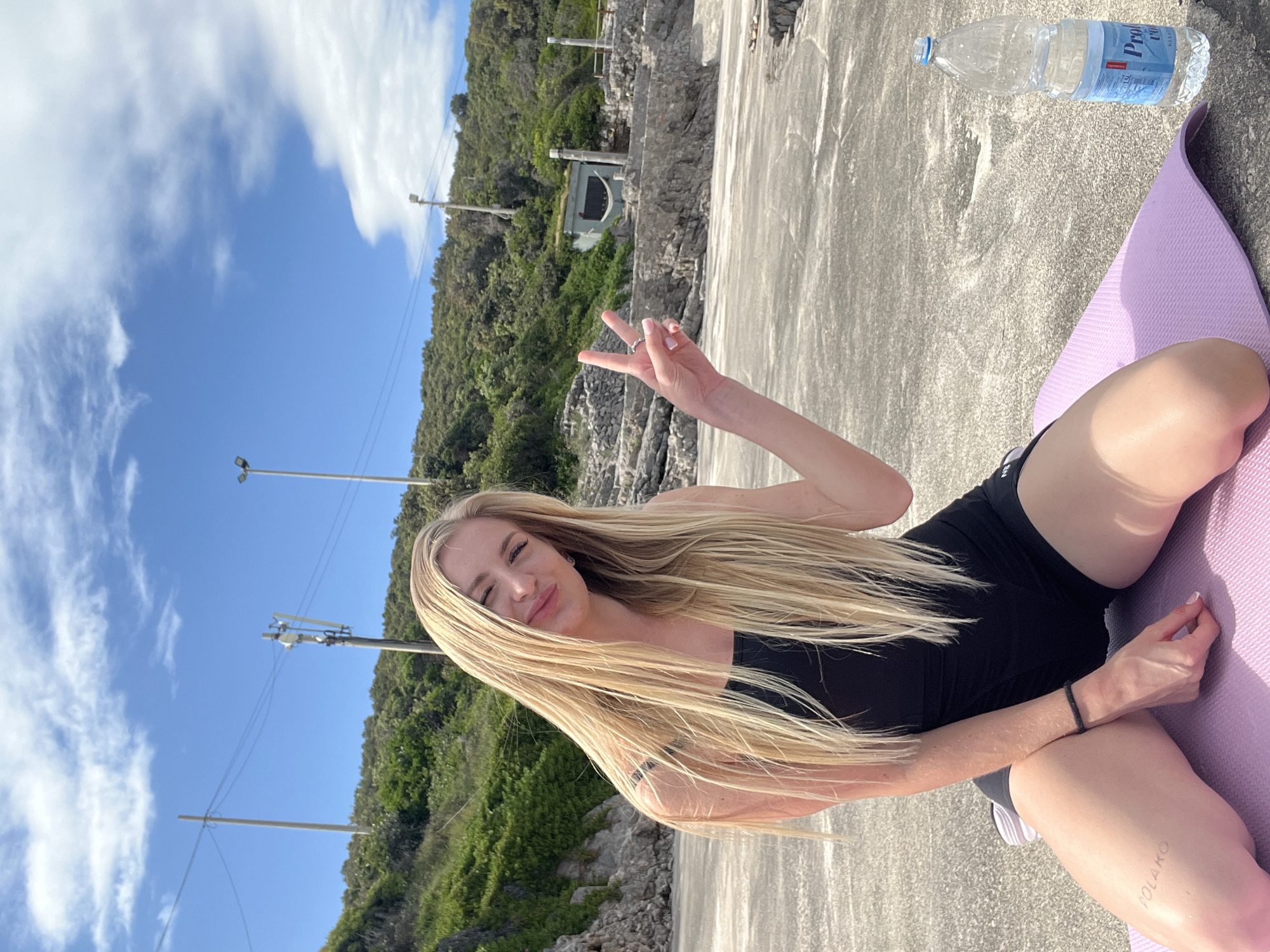 A young adult female with long blonde hair is sitting on a purple yoga mat on a gray concrete surface. She is wearing a black tank top and black shorts. She is holding up a hand with two fingers extended in a peace sign. A plastic water bottle labeled "Pro aqua" is near the upper right. Behind her is a green hillside with vegetation. Utility poles and a blue sky with white clouds are visible in the background. The location is in Prijevor, Montenegro.