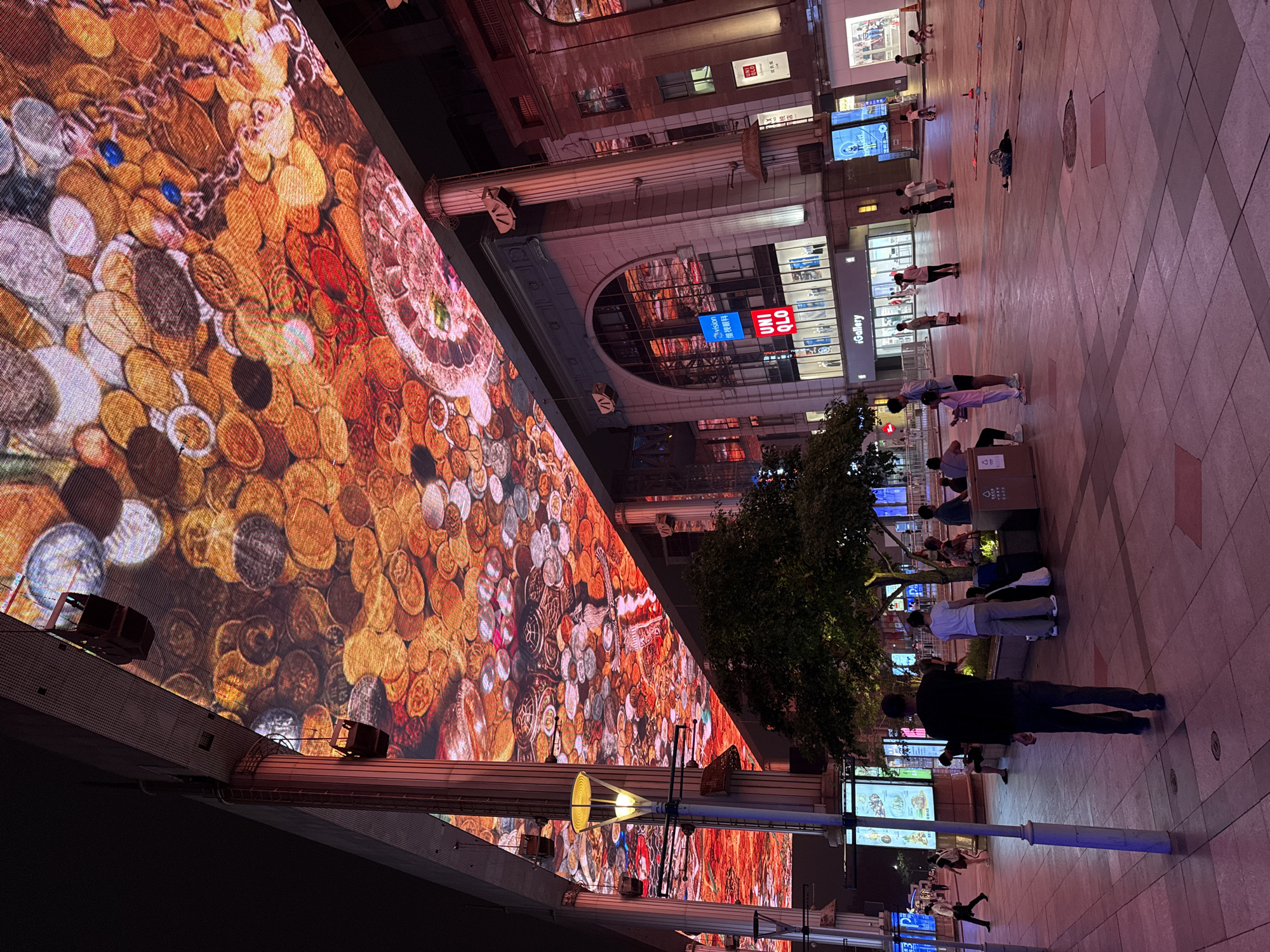 The image depicts a street scene at night in the Chaoyang District, China, featuring a large, vibrant LED screen displaying a pattern resembling coins and abstract colorful shapes. The screen is affixed to the underside of a covered walkway, casting a warm, reddish-orange light onto the pavement below. A building with large arched windows and illuminated signage for "UNIQLO" and "iGallery" is visible. Several people are present in the plaza, some walking, others standing or sitting. A lone figure in a black shirt and dark pants walks away from the camera towards the left. Other individuals are seen in groups and individually, with some children playing in the distance. Lampposts and trees are interspersed within the urban environment. The ground is paved with large square tiles. The overall atmosphere suggests a bustling commercial area at night.