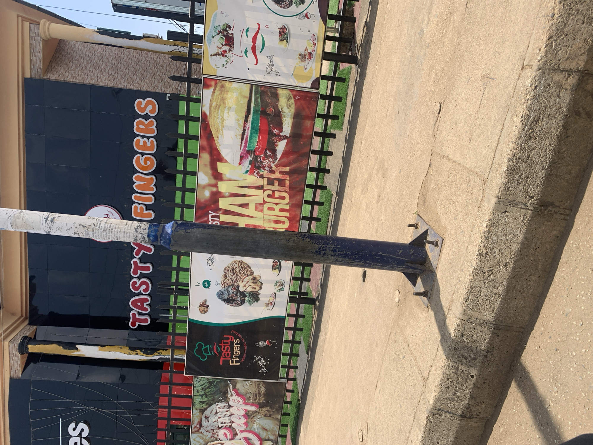 The image shows a street scene in Jos, Nigeria, featuring a restaurant named "Tasty Fingers". The restaurant's signage, including the name and menu items like "Iam Burger", is prominently displayed. The building has a black tile facade. A blue metal pole is visible in the foreground, obscuring part of the signage. The street is made of concrete. The overall environment appears to be urban.
