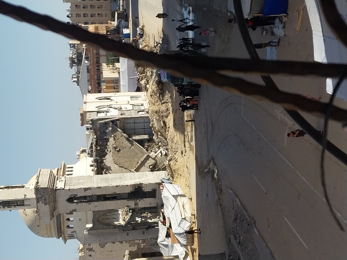 The image presents an elevated view of a devastated urban environment in Khan Yunis, Palestinian Territory, under a clear daylight sky. In the background, a large, light-colored building, identifiable as a mosque by its dome and minaret, exhibits severe structural damage. The minaret is partially collapsed, and large sections of the mosque's walls are missing, exposing interior spaces, with significant rubble accumulated at its base. To the left of the damaged mosque, several white tarpaulin tents are erected on sandy ground. A paved road with visible tire marks and scattered debris traverses the mid-ground. On the right side of the road, a group of approximately 20-30 individuals, including adults and children, is congregated near an unpaved area containing puddles; some are observed carrying plastic containers. Other individuals walk independently on the road and near the tents. A dark, out-of-focus metal railing or fence partially obscures the immediate foreground, indicating the photograph was taken from an elevated position.
