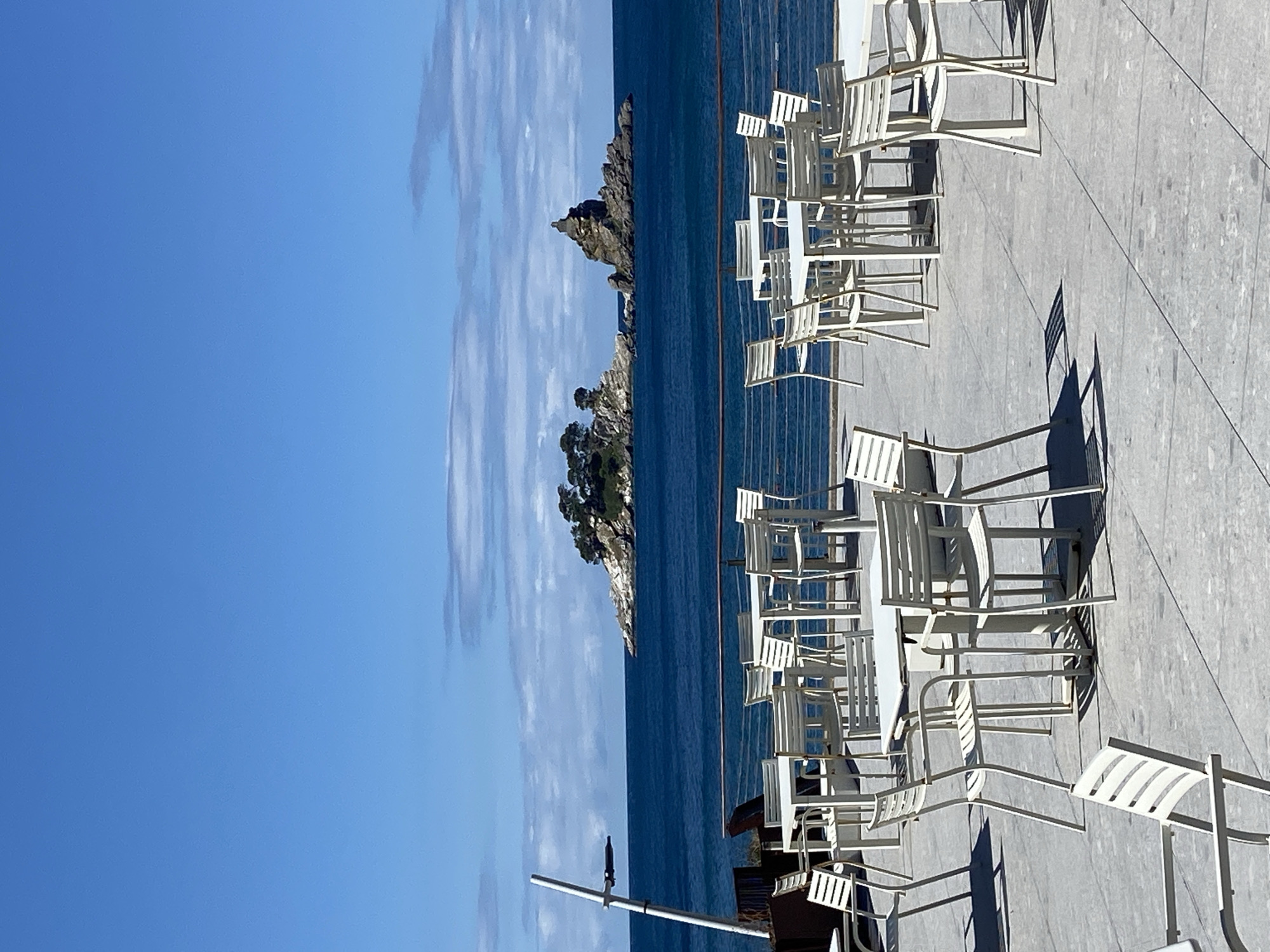 An outdoor seating area of a cafe or restaurant faces the Adriatic Sea in Brežine, Montenegro. White metal tables and chairs are arranged on a light-colored concrete patio. In the distance, a small rocky island dotted with trees and topped with a stone structure is visible in the deep blue sea under a bright blue sky with scattered clouds. A metal railing separates the patio from the water. A portion of a tall, thin white pole with a dark object attached is visible on the left side of the frame. The sun casts shadows from the tables and chairs onto the patio. No people are present in the scene.