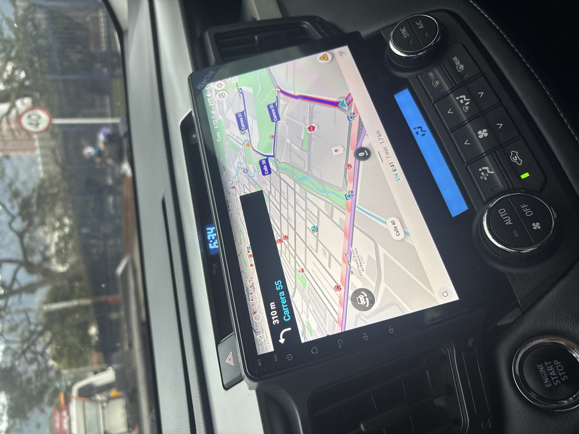 The image shows the interior of a car, focusing on the infotainment system displaying a navigation app. The map shows a route in Bello, Colombia. The destination is 310 meters away on Carrera 55. The ETA is 6:41 AM, with a travel time of 7 minutes and a distance of 1.7 kilometers. The current time is 6:32 AM, and the temperature is 21°C. The screen also displays nearby locations, including "AP Norte (N) EDS Texaco-Estrella del Renacimiento" and "Calle 40". The route on the map includes Carrera 57, Carrera 55, and Calle 38. The car's dashboard controls for the air conditioning and the engine start/stop button are visible.