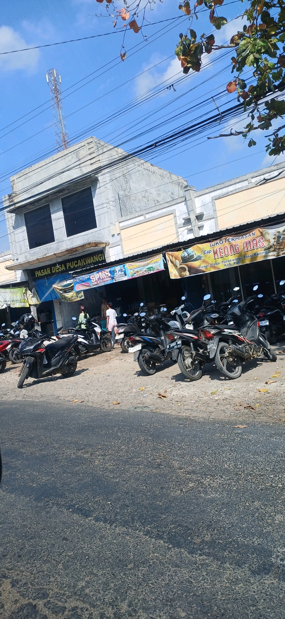 The image depicts a street scene in Pucakwangi, Indonesia, likely in front of a marketplace or commercial building. In the foreground, a paved road with a slightly textured surface is visible. Behind the road, a row of parked motorcycles of various makes and models are lined up. The motorcycles are predominantly dark in color, with some displaying license plates.
The background features a two-story building with a light-colored facade. The upper floor has two large, dark-tinted windows. The lower part of the building has awnings and signage. One prominent banner reads "PASAR DESA PUCAKWANGI," indicating the location as a village market. Another banner, featuring an image of a snail, states "TOKO PERHIASAN Cap KEONG MAS" (Jewelry Store, Golden Snail Brand). Other smaller signs and banners are also present.
Several people are visible in front of the building, some standing and interacting, while others are behind the counter or within the establishments. The people are dressed casually. The sky above is clear and blue with some scattered clouds, and utility wires are strung across the frame. In the distance, a tall telecommunications tower can be seen. The overall impression is a busy commercial area during daylight hours.