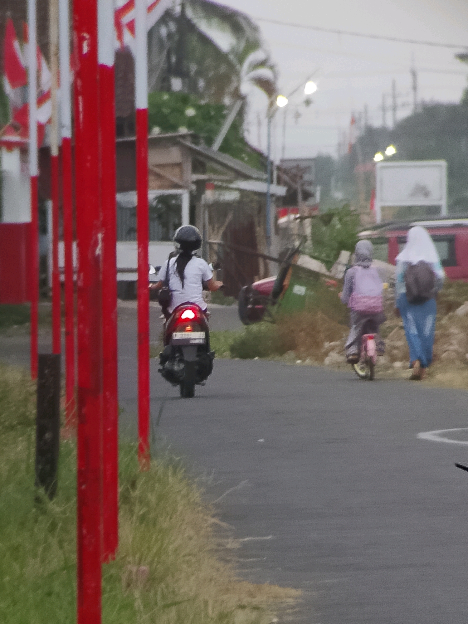In Jember, Indonesia, on a paved road, a person wearing a white shirt and black helmet rides a scooter. The scooter has a license plate and red taillights. Further down the road, another person on a pink bicycle, wearing a purple hoodie and carrying a pink bag, is observed. To the right of the cyclist, a person in a white hijab and blue skirt walks, carrying a backpack. The background features a red building, a green garbage bin, and an abundance of green vegetation. The scene is framed by red poles on the left side.
