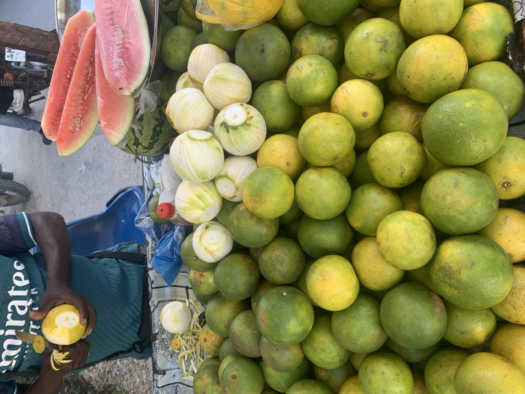 An overhead shot captures a vibrant fruit stall in Dar es-Salaam, Tanzania. The scene is dominated by a large assortment of fresh fruits, primarily round citrus-like fruits (likely oranges or pomelos) piled high, varying in shades of green and yellow, some partially peeled revealing white flesh. Several large, bright red watermelon slices, individually wrapped in clear plastic, are prominently displayed hanging on the upper left, with a whole watermelon visible beneath them.
On the left side of the frame, a person, likely a vendor, is engaged in peeling one of the yellow-green fruits with a small knife. Only their dark-skinned arms, hands, and torso are visible, wearing a dark blue/green striped shirt with the word "emirates" printed on it. Peels from the fruit are scattered on the surface beside their hands.
The setting appears to be an outdoor street market or roadside stall, indicated by partial views of a motorbike or bicycle and what looks like a street or dusty ground in the background. A blue plastic chair is visible behind the person, and a plastic bottle with a red cap sits near the fruit display. Bright, natural lighting suggests it is daytime. The image conveys a lively, everyday market scene focused on fresh produce.