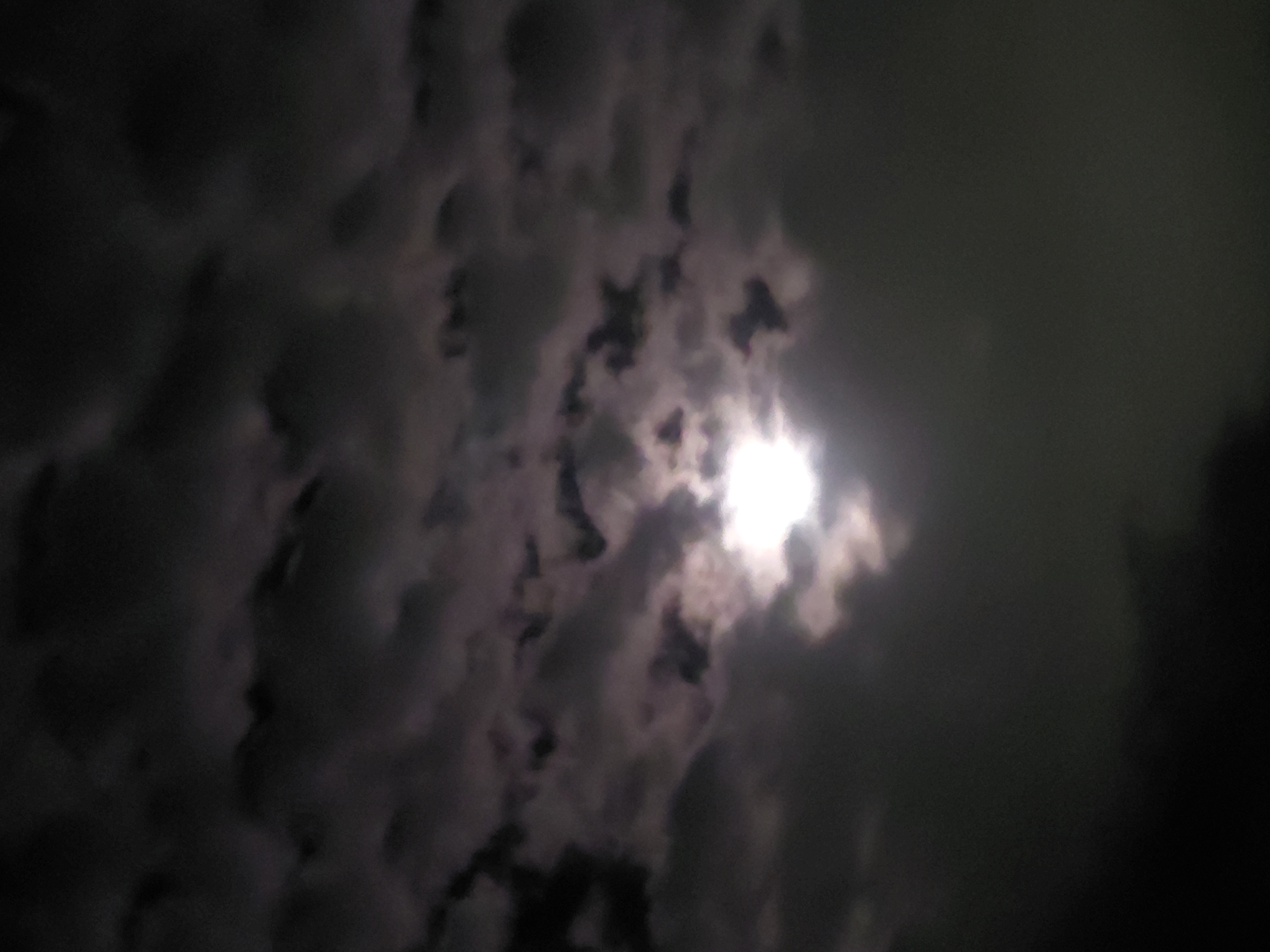 The image captures a dramatic nocturnal sky, heavily blanketed by a dense formation of clouds. A brilliant, glowing light source, strongly resembling the moon, pierces through an opening in the cloud cover, predominantly in the upper right-center of the frame. This intense light illuminates the edges and interiors of the surrounding clouds, creating a stark contrast between the luminous white and the deep, shadowy grays and blacks of the cloud masses.
The clouds themselves display varying textures and densities; some sections appear as dark, amorphous forms, while others are brightly outlined by the hidden light, revealing their intricate structures. The overall scene is devoid of people, man-made objects, or any discernible text. The image conveys a profound sense of mystery and serene power, characteristic of a cloudy night. Given the provided context, this natural phenomenon unfolds over Santa Ana, Venezuela. The time of day is clearly night, with the weather being overcast.