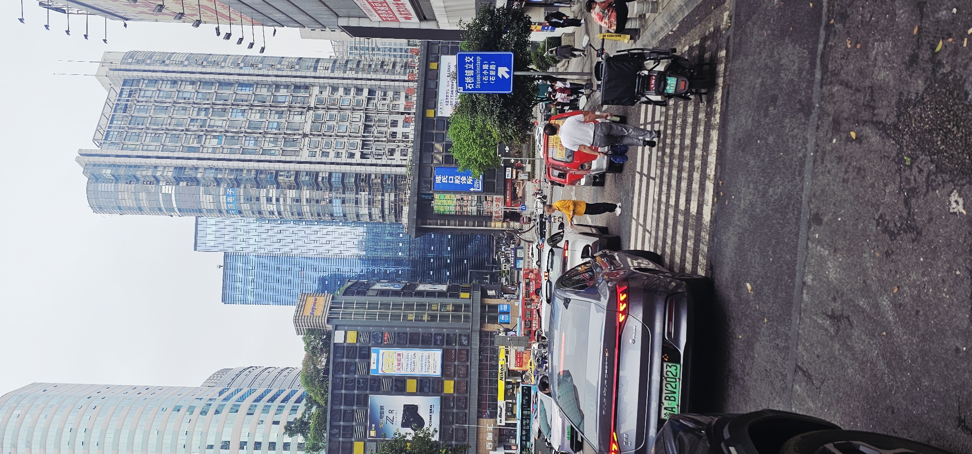 The image shows a street scene in Jiulongpo District, Chongqing, China. Tall buildings rise on either side of the street. People are seen crossing a crosswalk, along with several cars and a scooter. Street signs indicate the presence of the Shiqiaopu Interchange. Signs for "CINEMA" and "Nikon" are visible. The license plate of a gray sedan reads "A·BV2023".