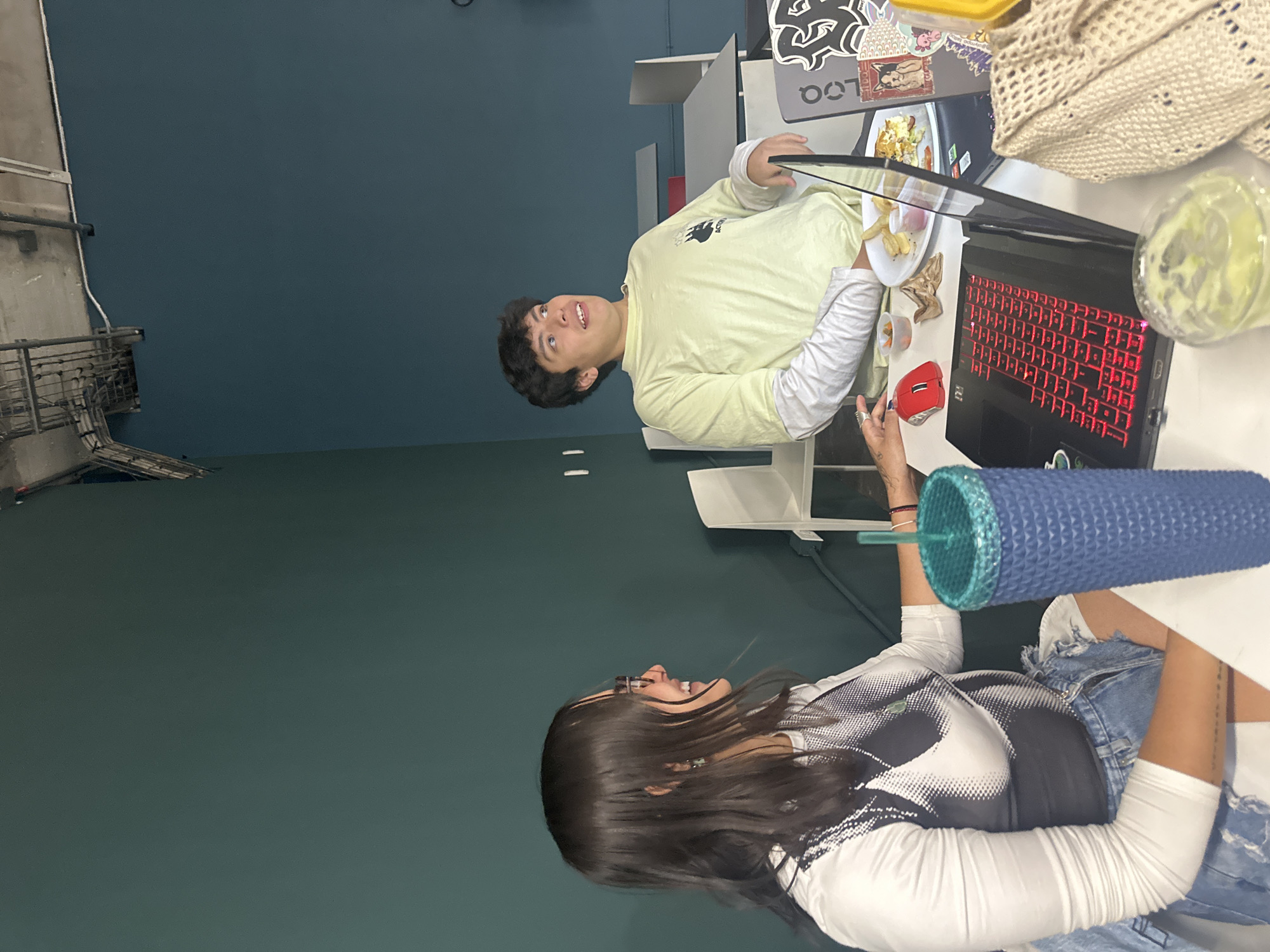 Two young adults are at a desk. One person wears a light yellow long-sleeve shirt and looks towards the camera. This person holds a black tablet and a plate of food. The second person wears a white and black long-sleeve shirt with jean shorts, holding a blue tumbler. They are both at a white desk with two laptops. One laptop is black with a red keyboard and a red mouse next to it. There is also a beverage and a crochet bag. The location has green and blue walls. Through a window, an exterior showing a metal structure suggests an industrial setting. The location is in Mexico City, Mexico.