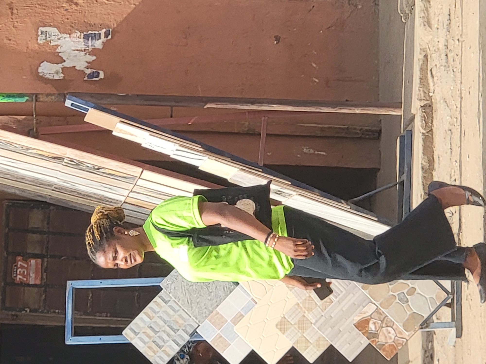 The image depicts a woman standing outdoors, likely at a building materials store or market in Maiburiji, Nigeria, surrounded by various floor and wall tiles.
The subject is a woman with light brown skin and dreadlocks or braids styled into a bun. She is wearing a bright lime green short-sleeved top, dark trousers, and dark flat shoes. A black shoulder bag is worn across her body, and she has decorative earrings and multiple bracelets on her right wrist. She is holding a dark-colored smartphone in her left hand, looking downwards at the tiles or her device. Her expression appears neutral or slightly smiling.
The scene features numerous patterned tiles displayed on low stands in the foreground and midground. These tiles exhibit a variety of designs, including geometric patterns, textured surfaces, and stone-like finishes. Behind the woman, stacks of wooden planks or other construction materials are visible. The background consists of textured walls: a reddish-brown wall with torn paper advertisements and a darker brown wall composed of small square tiles.
Notable details include a small red sign with the number "737" prominently displayed in white on the tiled wall. The lighting is bright and natural, indicating daytime. The environment suggests an open-air commercial space for construction supplies.