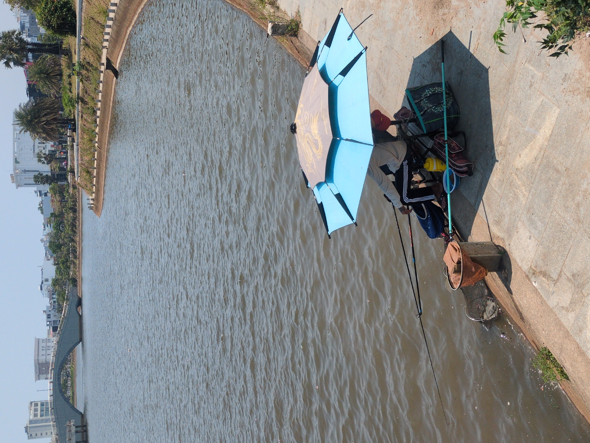 The photograph captures a fishing scene in Tuy Hòa, Vietnam. An individual is sitting on a foldable chair beneath a blue and gold umbrella, fishing on the edge of a water body. Fishing gear, including a rod and a net, are visible. The surroundings consist of a stone embankment and water with ripples. Buildings and greenery are seen in the background, suggesting an urban environment.