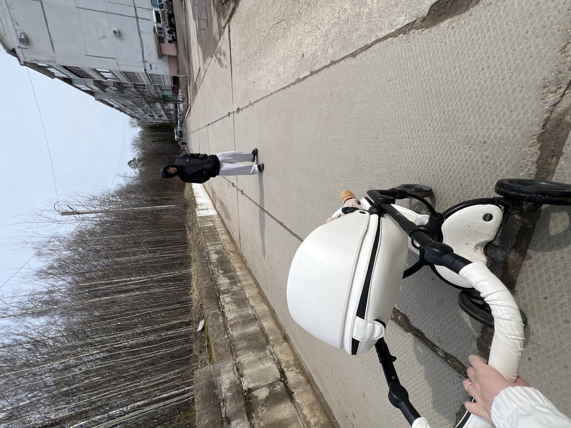 The image shows a person pushing a white stroller on a concrete sidewalk in Noviy, Russia. A person wearing a black jacket and light gray pants walks ahead on the sidewalk. To the left is a row of bare trees, and further back is a multistory apartment building. The sidewalk is cracked in places. The person pushing the stroller is visible from the arm up, wearing a white long-sleeved shirt.