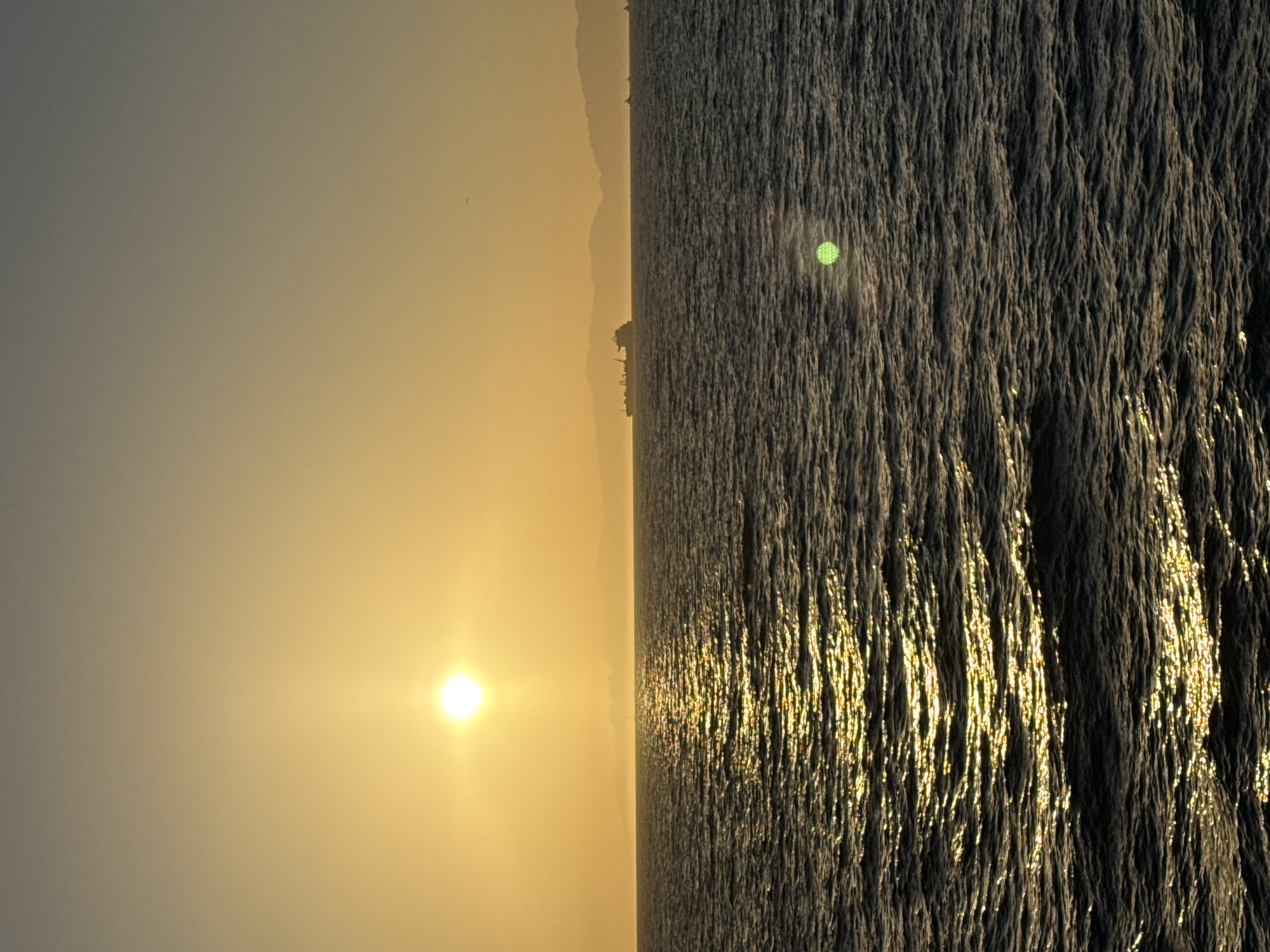 A wide-angle vertical photograph captures a serene ocean sunset. The sun, a bright, luminous orb, is positioned in the upper left quadrant, casting a golden, shimmering path across the rippling water. The sea is dark, with waves creating a textured surface reflecting the sunlight. In the distance, a faint, hazy silhouette of mountains or hills forms the horizon line. A large cargo ship and several smaller vessels are visible on the water in the mid-ground. The sky transitions from a warm yellow near the horizon to a muted grey at the top of the frame. The scene appears to be set at sea, likely offshore from a coastal area, and depicts a natural environmental landscape devoid of any urban elements that would specifically identify it as Manhattan Beach, United States of America. There are no people or specific actions depicted in the image.