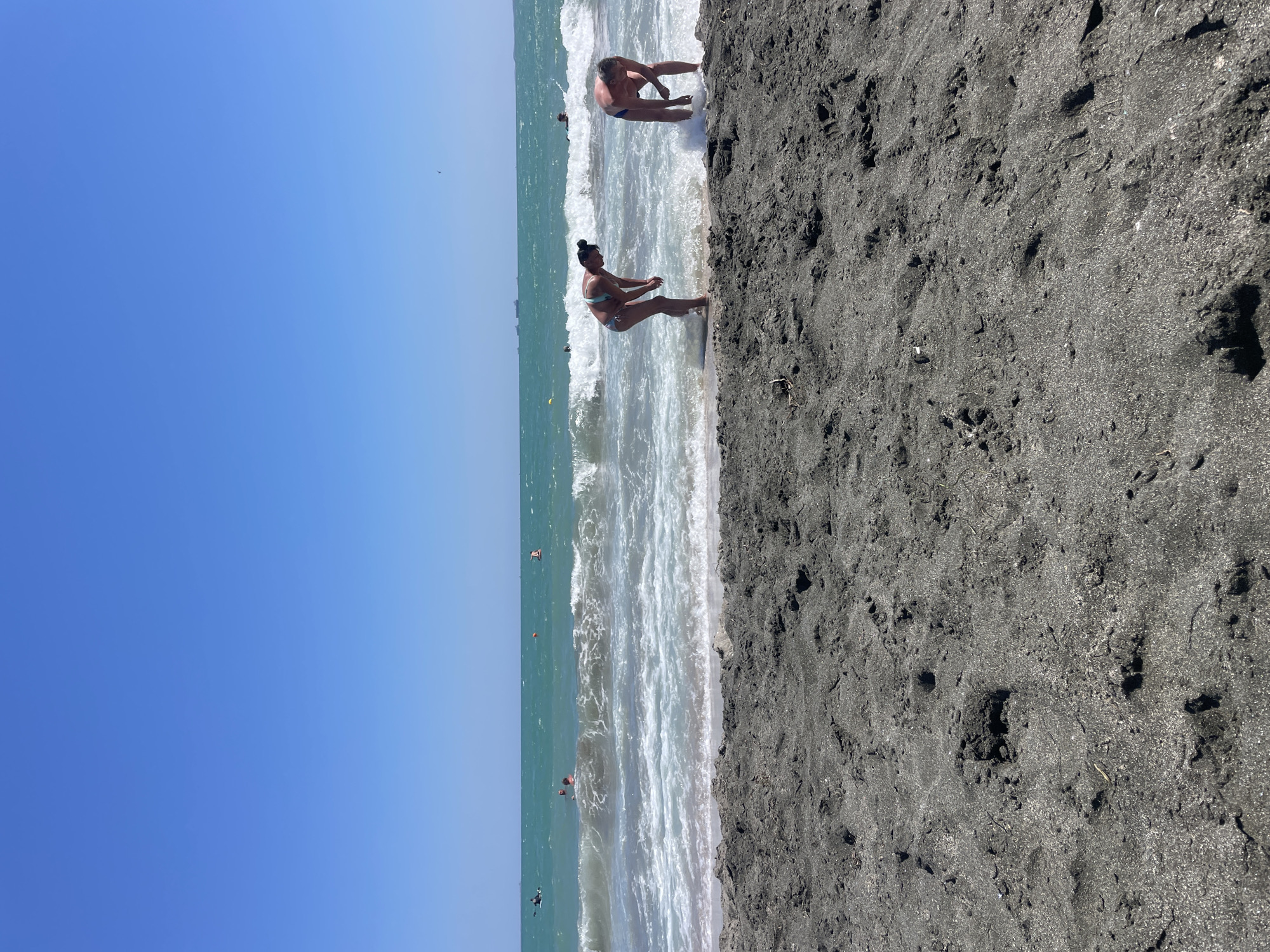 This image captures a sunny day at the beach in Burgas, Bulgaria, with the Black Sea stretching out under a clear blue sky. The foreground is dominated by dark, coarse sand, showing footprints and small indentations. In the midground, two individuals are positioned at the water's edge. To the left, a woman wearing a light blue bikini bends forward, seemingly interacting with the shallow water. To the right, a man in blue swim trunks also bends down, with waves splashing around his legs. In the background, the turquoise sea churns with white-capped waves. Several other people are visible in the water further out, some swimming and others on flotation devices. Distant cargo ships can be seen on the horizon, suggesting maritime activity. The overall scene depicts a typical summer day at a seaside resort.