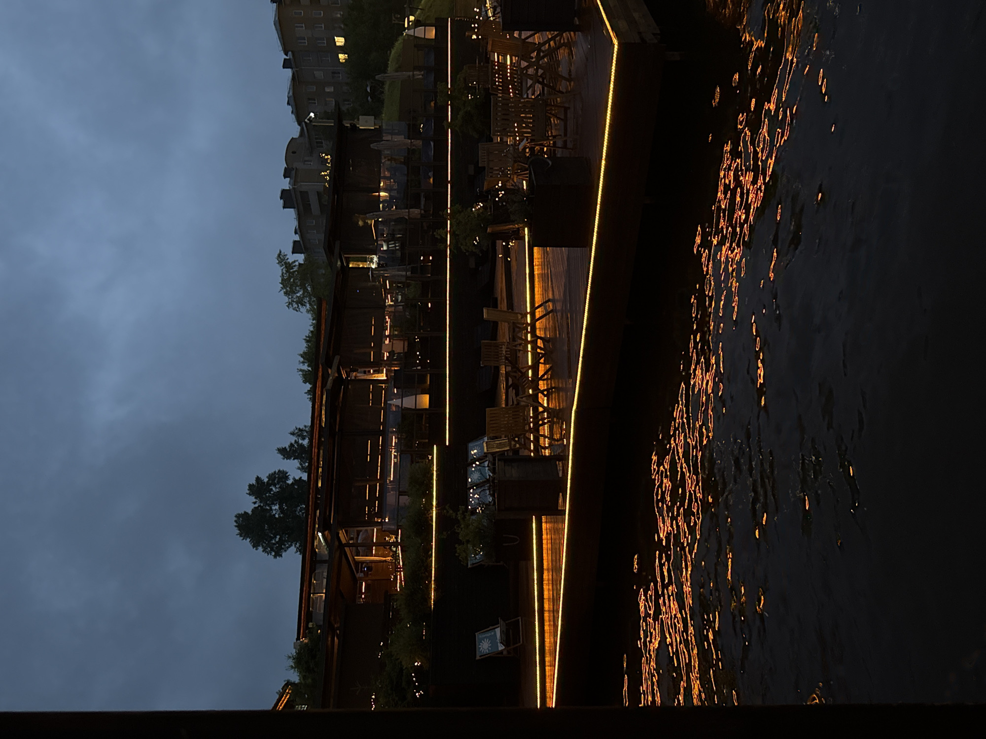 This image presents an outdoor multi-tiered dining or leisure complex situated on a body of water in Votkinsk, Russia, captured during the evening or night. The sky is dark and overcast, with the silhouettes of deciduous trees and multi-story residential buildings visible in the distance along the horizon. Several windows in the distant buildings show illuminated warm light.
The central feature is a wooden deck system extending over the water, brightly illuminated by continuous, warm-toned LED strip lighting along its perimeters and structural elements. An upper-level structure, likely a restaurant or bar, features glass walls, a dark roof, and visible interior lighting with tables and chairs. Below this, lower deck sections contain numerous unoccuppied wooden tables and chairs arranged for outdoor seating. Dark-colored planter boxes containing green foliage are interspersed among the seating areas. Two light-colored lounge chairs with patterned fabric are visible on one of the lower deck sections to the left. No people are present within the observable areas of the complex. The foreground comprises a dark body of water, its surface actively reflecting the warm, orange-yellow artificial lights from the deck and structures above as elongated, undulating patterns, indicative of minor surface ripples.
