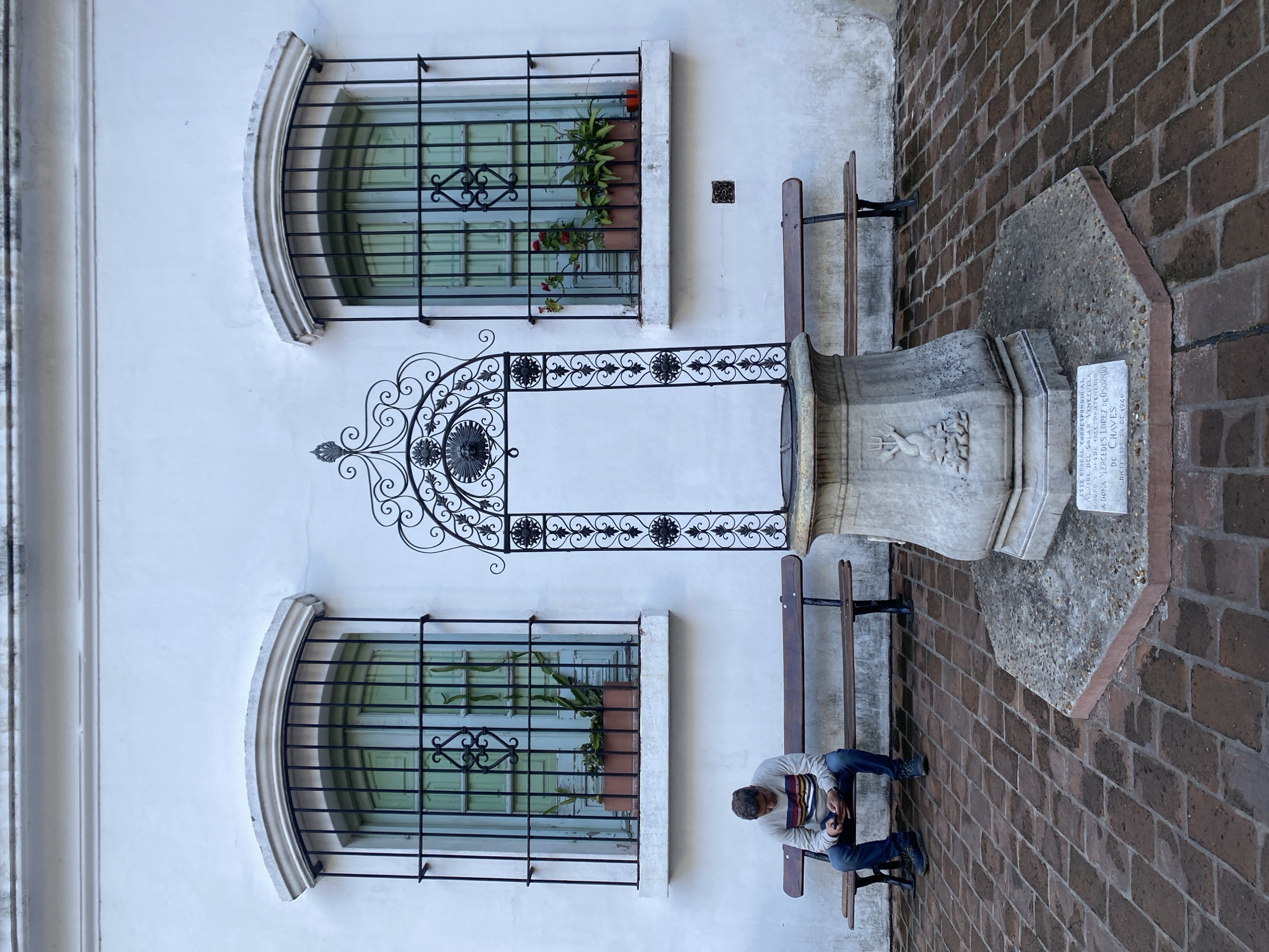 A man sits on a wooden bench beside a historic stone well in Buenos Aires, Argentina. The well, likely associated with the 'solar Venezuela' referenced on the plaque, is surrounded by brick paving and flanked by two benches. The wall behind the well features two arched windows with black wrought iron grilles, each containing a planter with greenery. An ornate wrought iron decoration hangs between the windows. The man is looking down, possibly at a mobile phone. The photo is taken at a slight angle, giving a tilted perspective.