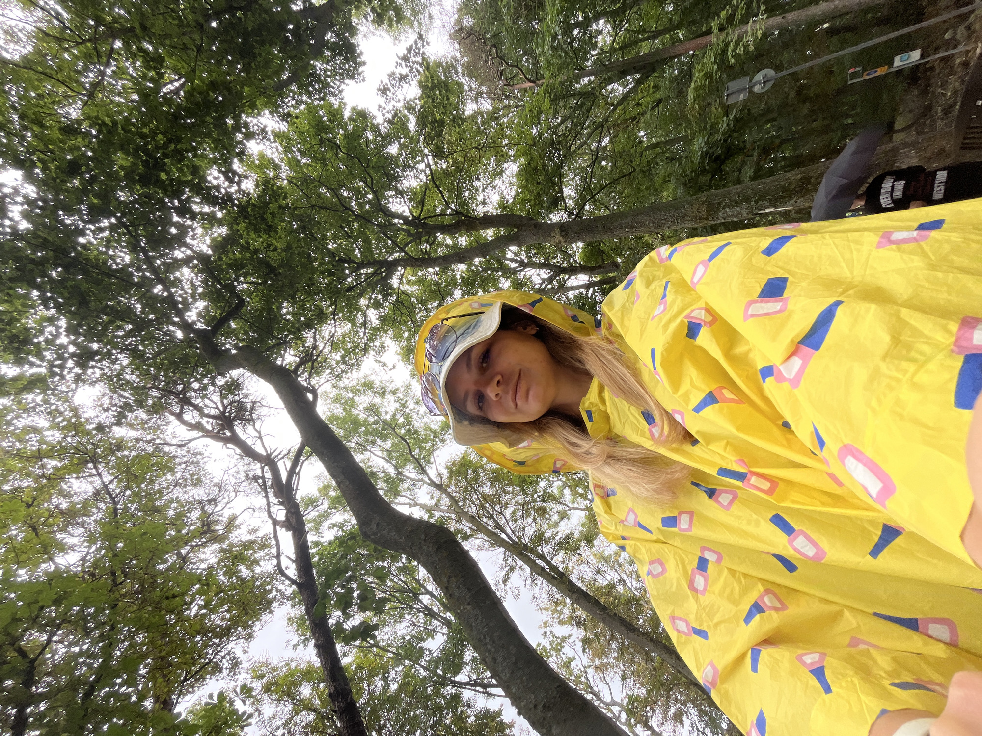 A medium-shot selfie taken from a low angle features a blonde woman wearing a yellow raincoat with a nail polish bottle pattern and a white bucket hat. She is looking at the camera with a neutral expression. The background consists of a forest with lush green trees and a cloudy sky. The woman appears to be in an outdoor setting, possibly a park or hiking trail. A road sign and another person holding an umbrella can be seen in the background on the right side of the image. The overall scene suggests a rainy or overcast day in a natural environment.