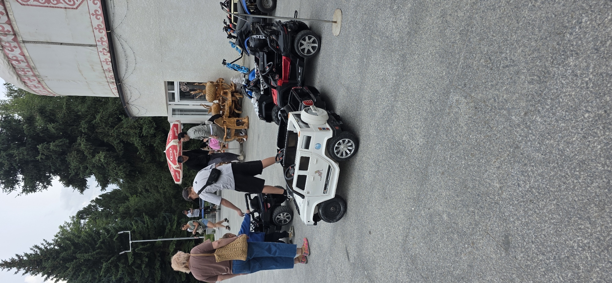 In Bulan-Segettu, Kyrgyzstan, a scene unfolds in front of a white building with a traditional yurt-like structure on its roof. The ground is paved with grey asphalt. Several electric toy cars are parked on the asphalt, including a white Hummer-style vehicle with a spare tire on the back, and a red and black vehicle. In the background, a red and white umbrella provides shade over a group of people gathered at wooden tables and chairs. A white pole with lights stands to the left, near a dense line of tall pine trees. Two women are standing in the foreground, one with a straw bag over her shoulder, and another woman is in the middle ground looking at her phone. A person in a grey t-shirt and black shorts is bent over one of the toy cars, interacting with a child. Other individuals are visible in the background, some standing and some sitting.
