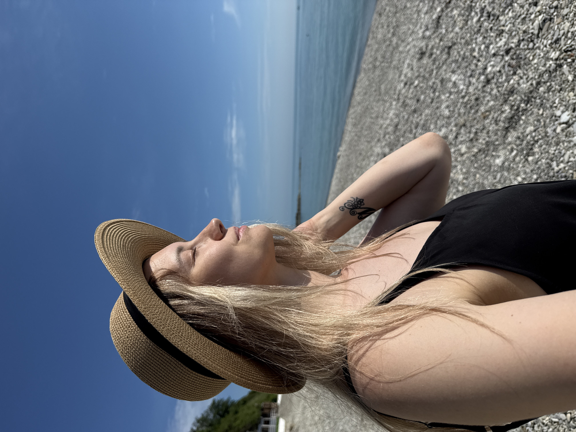 A woman with long blonde hair, wearing a straw hat and a black dress, stands on a pebble beach. She is looking to the right with her eyes closed and her hand on her neck. The sea is visible in the background, with a clear blue sky above. The location is Nebug, Russia.