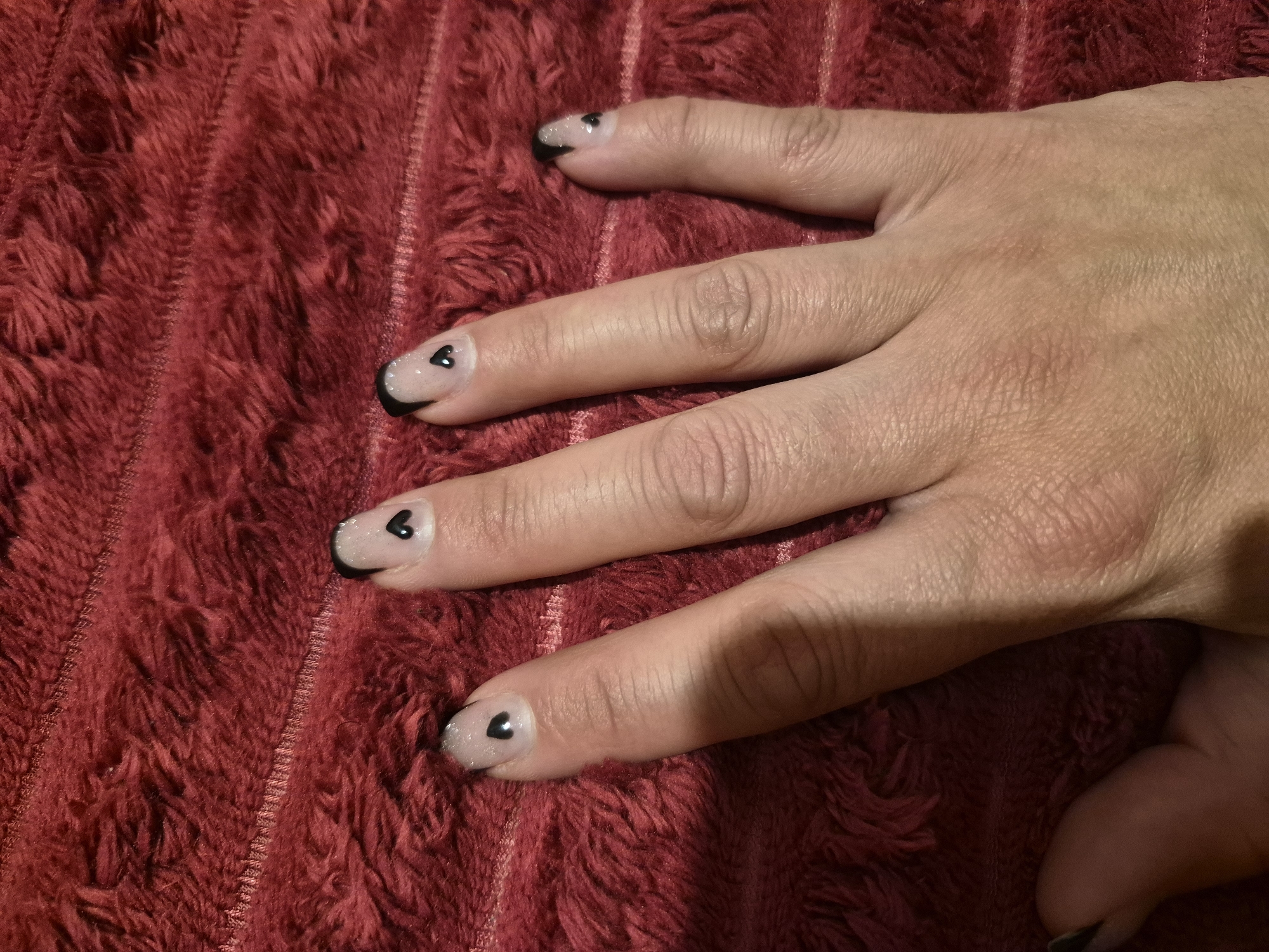 The image shows a close-up of a person's left hand with black heart-shaped nail art on a light, sheer base coat. The background is a textured, red fabric. There is no identifiable information about the location in Sandwell, United Kingdom, as the image only focuses on the hand and the fabric.