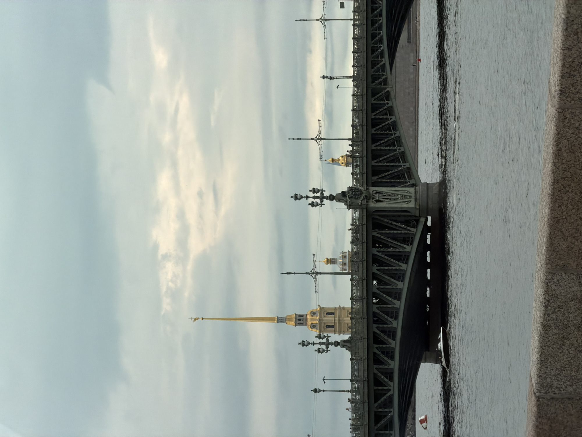The image captures a view of a bridge spanning a body of water under a cloudy sky. The bridge is a substantial structure with intricate metalwork and decorative lampposts. On the far bank of the water, a tall, slender, golden-spired building, likely a church or cathedral, is prominently visible, with smaller golden domes also present. Several figures can be seen walking on the bridge. A small boat is navigating the water beneath the bridge. The foreground features a textured stone embankment. The scene appears to be in St. Petersburg, Russia, with the golden spire likely belonging to the Peter and Paul Fortress.