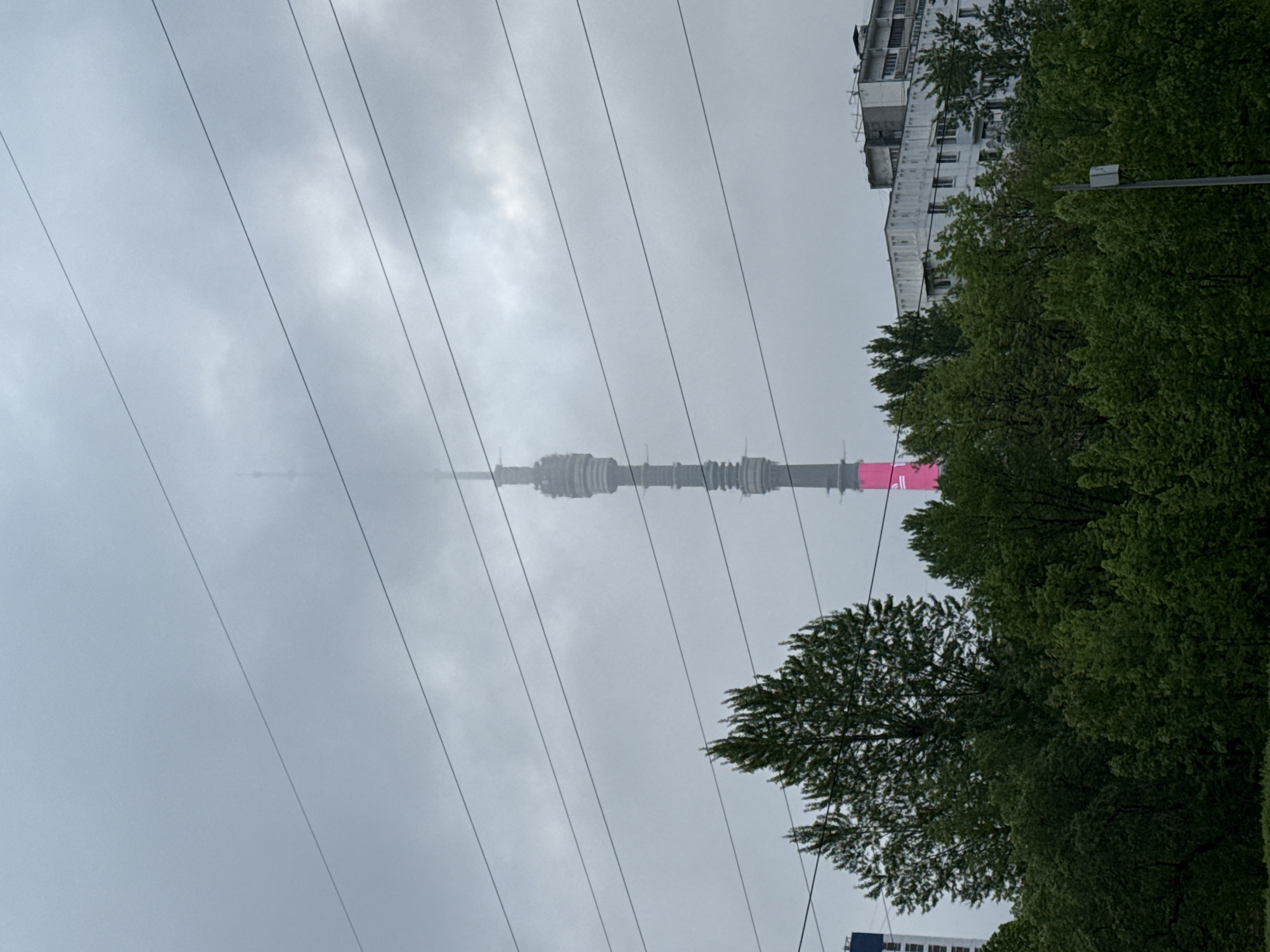 The image depicts the Ostankino Tower in Moscow, Russia, partially obscured by a low cloud ceiling. The tower, grey with some pink, rises above tree lines. Power lines run across the foreground of the image. A multi-story building can be seen to the right of the tower. The sky is overcast. The specific location in Khimki cannot be determined from the image.