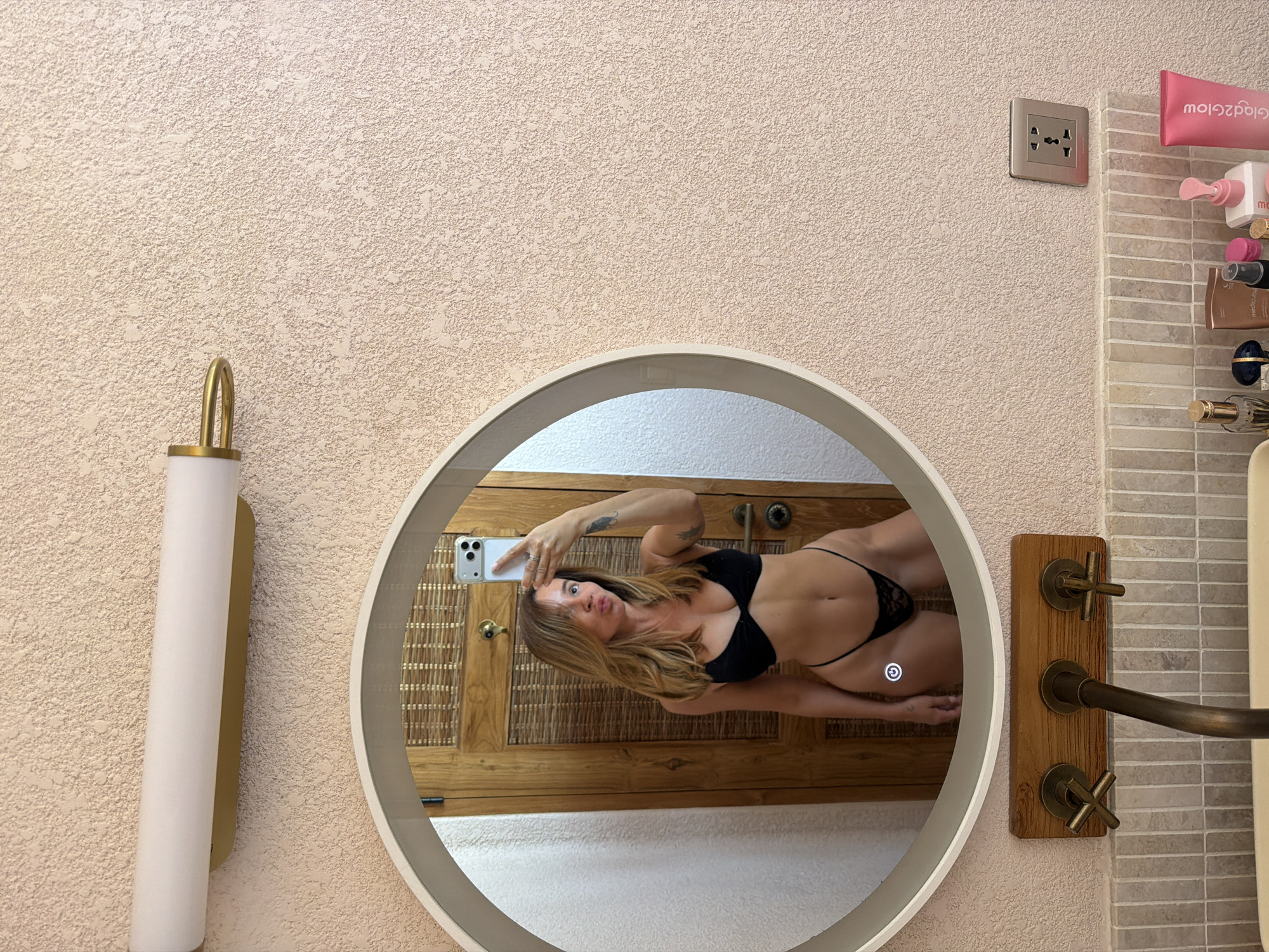 The image shows a woman in a black bikini taking a selfie in a mirror. She is standing in what appears to be a bathroom. The mirror is circular and has a light around its edge. Behind her, there is a wooden door with a woven texture. In front of the mirror, there is a sink with brass faucets. The wall is textured and painted a light pink color. On a shelf below the mirror, there are several bottles of toiletries, including a pink tube labeled "Glad2Glow." The scene appears to be in a residential setting.