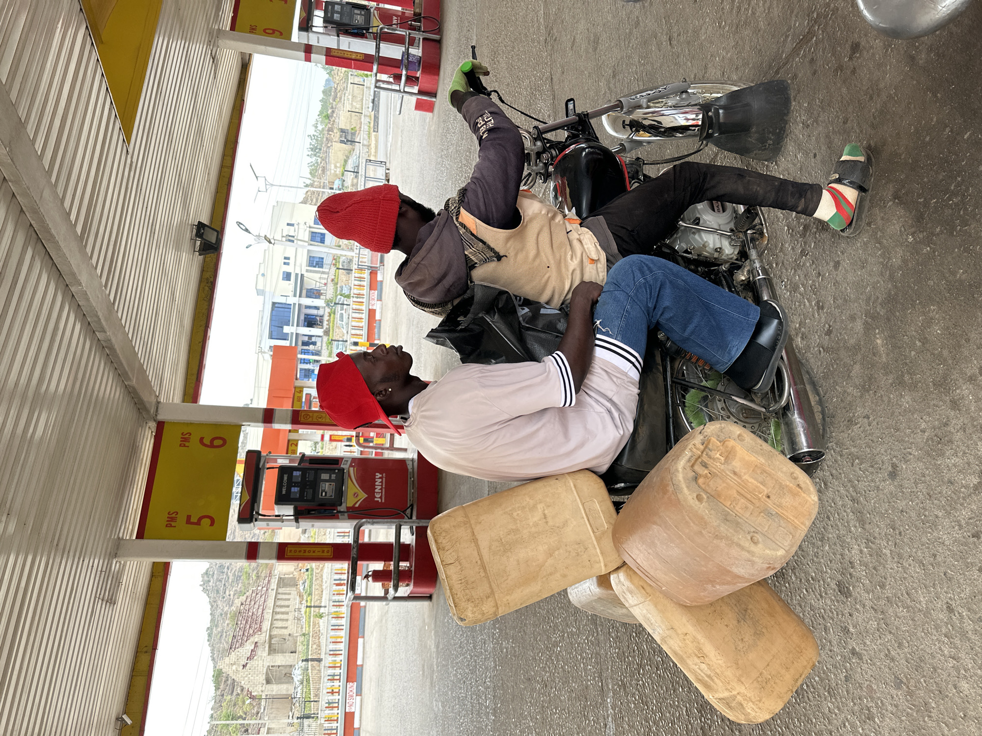 This image captures a scene at a gas station in Jos, Nigeria, under bright daylight. Two men are seated on a black and chrome motorcycle, positioned in front of fuel pumps. Both men wear red knit caps. The man closer to the viewer is in a light-colored t-shirt with black and white striped short sleeves and blue jeans, with black dress shoes. He is looking upwards, towards the man in front of him. The second man, facing away from the camera, wears a dark long-sleeved top, a light-colored vest, and a checkered scarf draped over his shoulder. His feet are visible, one wearing a green sock with red stripes and a black sandal.
Stacked on the concrete ground beside the motorcycle and the first man are several large, light brown plastic jerry cans, suggesting they are purchasing or transporting fuel in bulk. In the background, visible through the open structure of the gas station, are developing urban areas with buildings under construction and hilly terrain.
Notable details include the gas station canopy and pumps. Yellow signs indicating "PMS 5" and "PMS 6" are attached to the structural poles. A red fuel pump branded "JENNY" displays "WELCOME" and "92" on its screen. Multiple red and white striped poles are visible, bearing "NO SMOKING" text. The ground is grey concrete. The overall atmosphere suggests a routine stop for fuel, possibly for commercial purposes given the jerry cans.