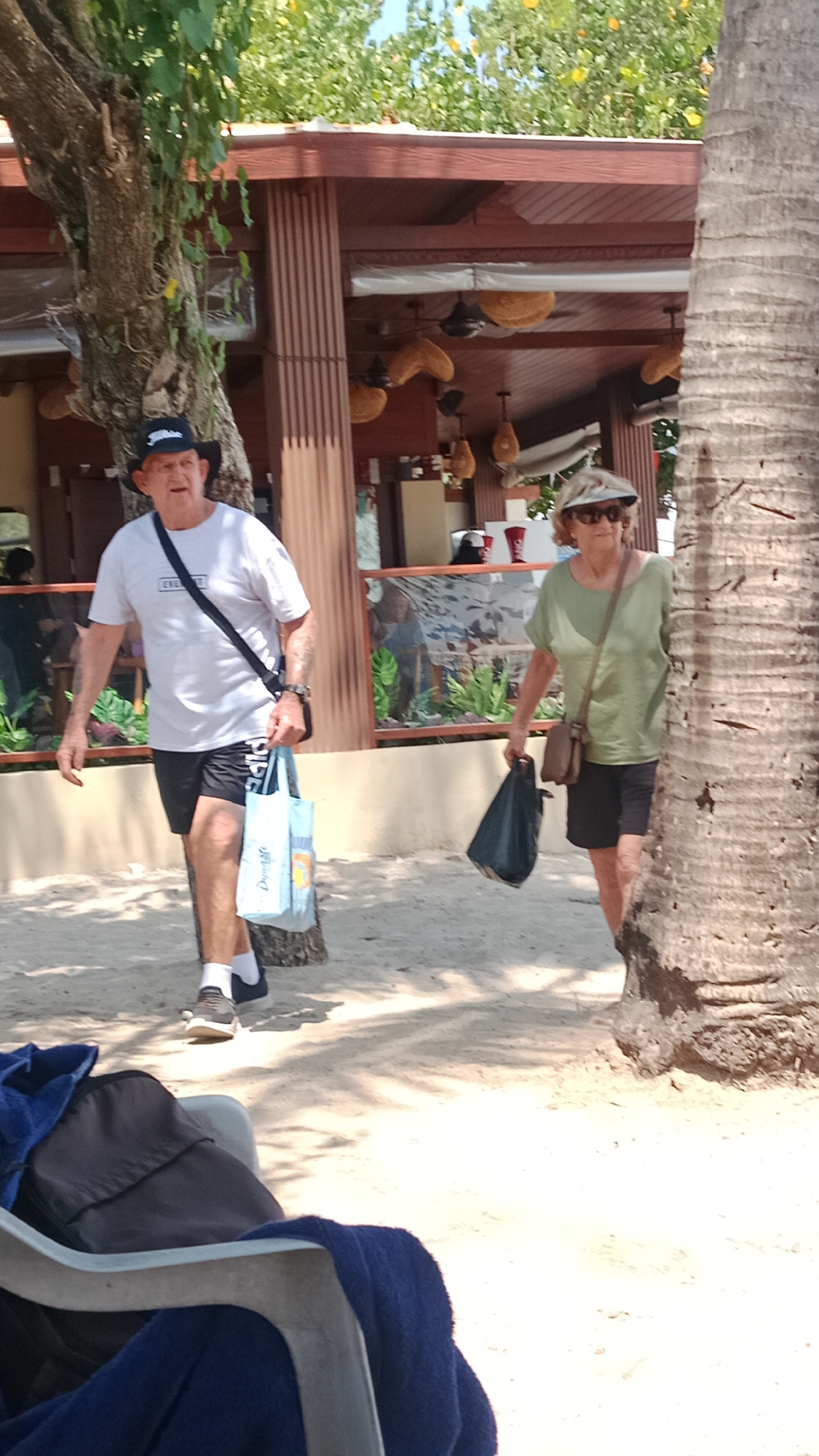 In Kuta, Indonesia, a man and a woman are walking on a sandy path. The man, on the left, wears a white t-shirt, black shorts, a black baseball cap, and dark shoes with white socks. He carries a black crossbody bag and a light blue shopping bag. The woman, on the right, has on a light green t-shirt, dark shorts, a visor, and carries a brown shoulder bag and a dark plastic bag. They are walking past a building with a covered outdoor seating area, featuring wicker light fixtures. Tropical trees with green foliage and yellow flowers are visible in the background. The ground is covered in sand, and the scene is illuminated by sunlight, casting shadows. A portion of a beach chair with blue towels is in the foreground.