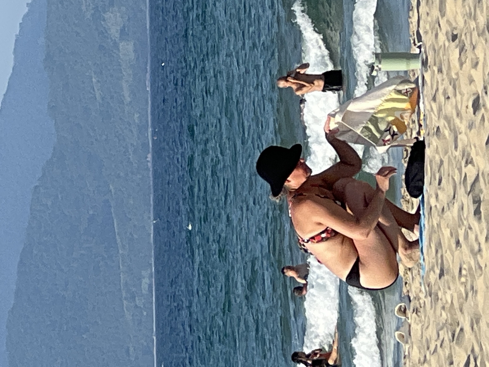 This media file captures a beach scene in Đà Nẵng, Vietnam. The primary subjects are several people engaged in various activities on the sandy shore and in the shallow water. In the foreground, a woman wearing a black hat and a patterned bikini is seated on a blue mat. She is positioned facing towards the ocean, with her body angled to the side. Another person is visible behind her, also seated on the mat and holding a piece of fabric.
Further back, other individuals are present. One person is standing in the water, with their arms raised, possibly playing or interacting with the waves. Another figure is partially visible in the water. In the distance, a sandy beach stretches out, meeting the ocean. The water is a deep blue, with white waves crashing along the shore. The background features a hazy, mountainous landscape.
The time of day appears to be sunny, with clear skies indicated by the bright lighting. The scene conveys a relaxed atmosphere, typical of a day at the beach. There is no visible text in the media file. The presence of the mountains and the style of the beach are consistent with the location context of Đà Nẵng, Vietnam.