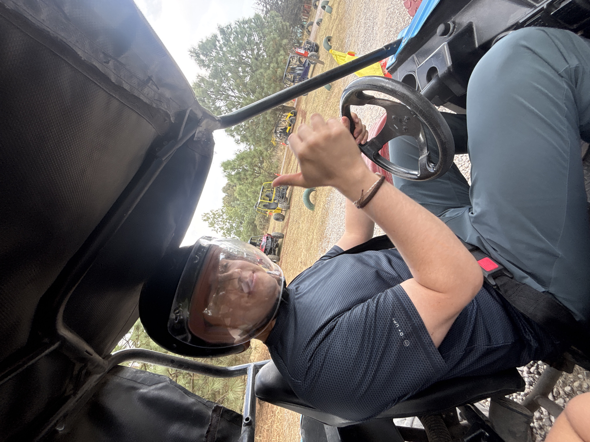 A person wearing a helmet with a clear visor is seated in a go-kart. The person is holding the steering wheel with their left hand and wearing a bracelet on their wrist. A seatbelt is fastened across their torso. The go-kart has a black canvas roof and visible frame, and other go-karts are visible in the background. The location is San Juan Arcos Ojo de Agua, Mexico. The ground appears to be covered in brown mulch or pine needles. The sky is overcast.