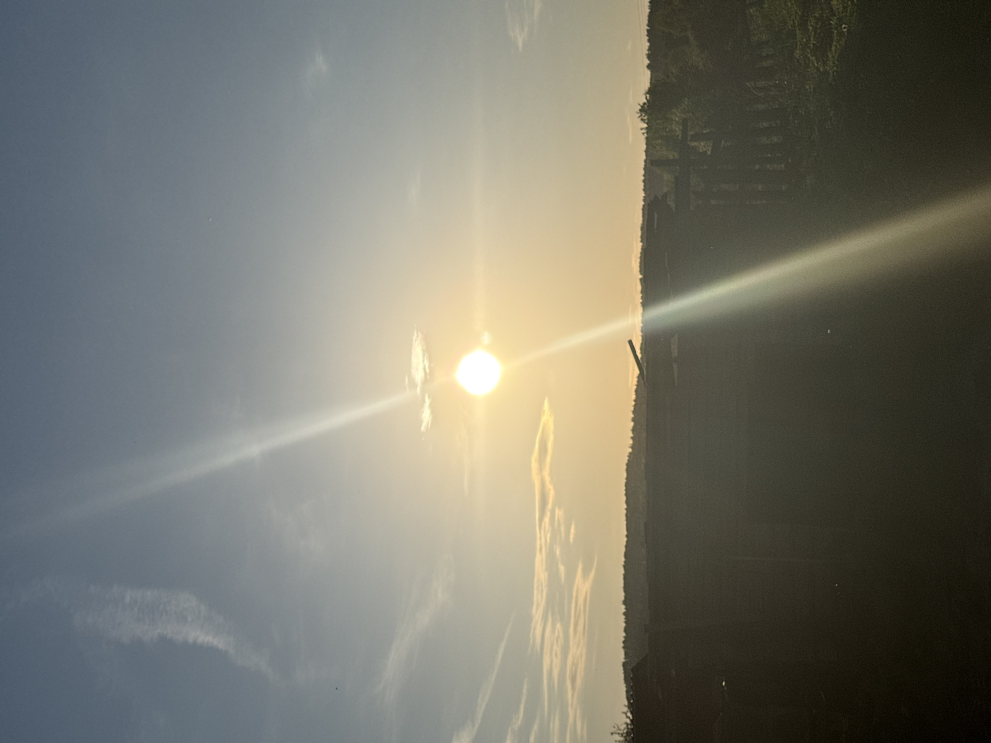 The image depicts a bright sun low in the sky over Nizhnyaya Chermoda, Russia. The location is identifiable by the typical Russian rural scene with wooden fence and scattered shrubs that is heavily obscured. The time of day appears to be either sunrise or sunset. The sky is predominantly blue with wispy clouds, transitioning to a golden hue near the horizon where the sun is positioned. A strong lens flare emanates from the sun. The right side of the image shows a silhouette of vegetation and what appears to be a wooden fence or structure. The details are largely obscured due to the heavy contrast and darkness in that area.