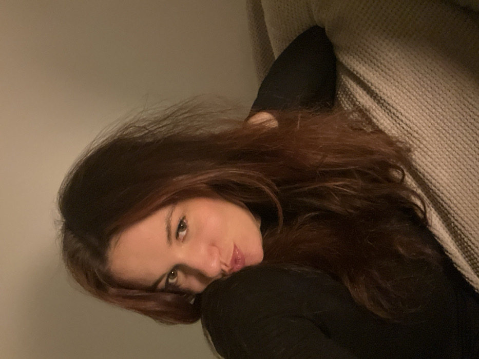 A woman with long, dark brown hair rests on a beige textured couch. She wears a black, long-sleeved shirt. Her head is tilted toward the camera, and she has a slight pout. The background is a plain, light gray wall. The lighting is soft, creating shadows that emphasize the contours of her face. Her gaze is direct, and she appears relaxed.