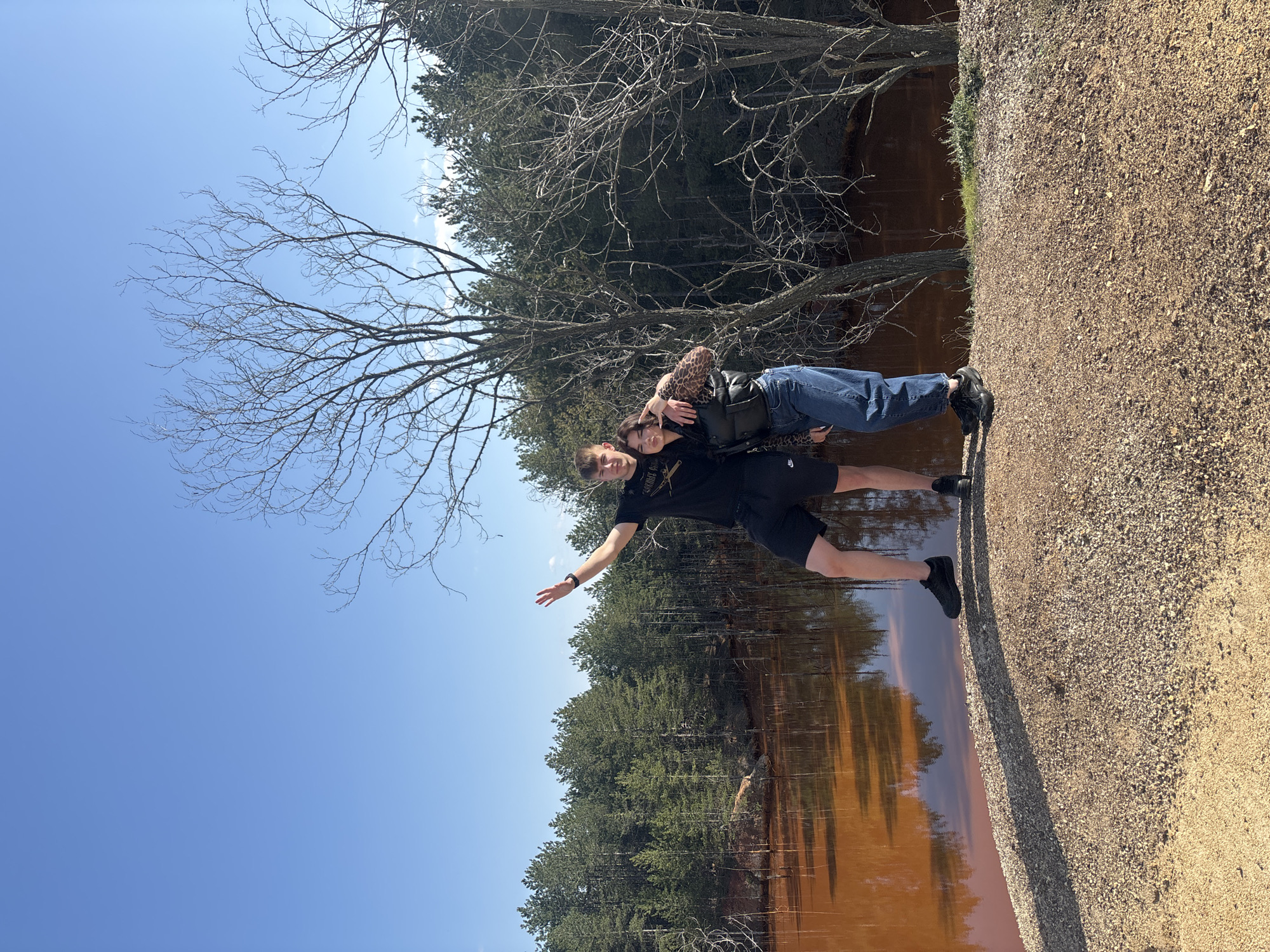 Two people stand near a body of water, likely a lake, in Elshitsa, Bulgaria. One person is wearing blue jeans, a vest, and a leopard print shirt, while the other is wearing black shorts and a black shirt. The water appears brown or reddish, possibly due to mineral content. The area includes trees and gravel. One of the people is extending their arm out to the side. The sky is clear and blue.