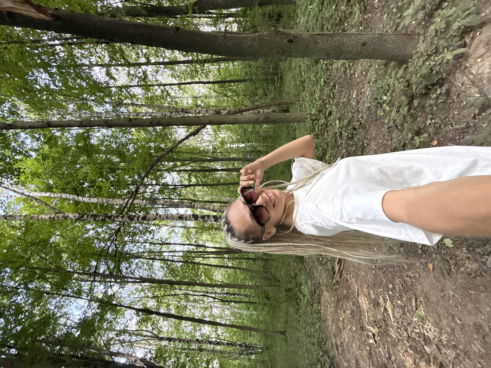 A young woman with blonde braids and sunglasses smiles at the camera while adjusting her sunglasses. She is wearing a white t-shirt and standing in a forest in Rusanovo, Russia. The forest is filled with tall trees and green foliage, and the ground is covered in dirt and fallen leaves. The woman is holding the camera out in front of her, taking a selfie. The image appears to have been taken during the daytime.