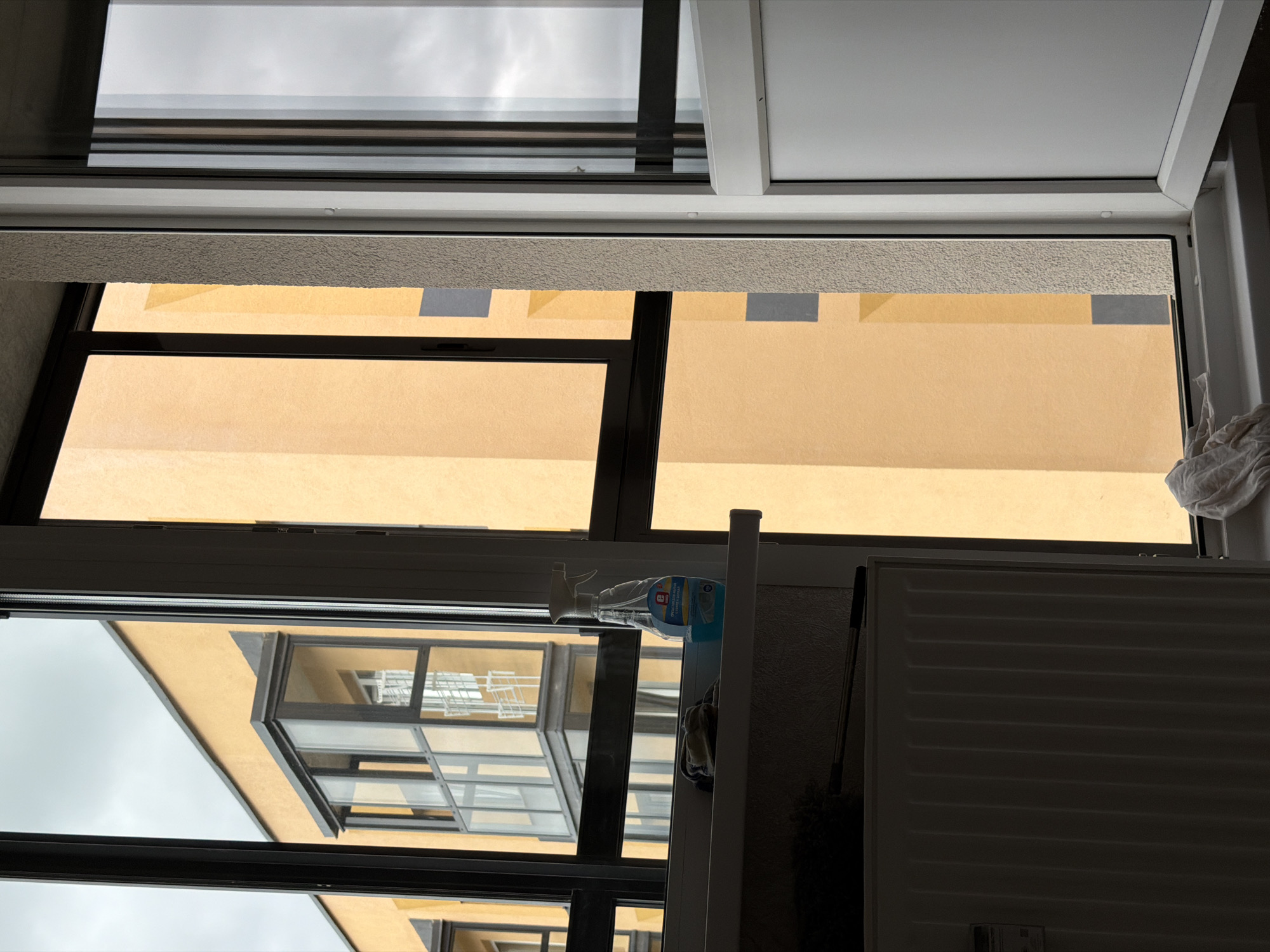The image captures a view through a modern window with black frames. The exterior reveals a building with a bright orange facade and a balcony with glass panels. The sky is overcast. On the interior windowsill, a bottle of cleaning spray and a rag are visible. Below the windowsill, a radiator is partially visible, shrouded in shadow, with a cleaning mop leaning against it. The scene appears to be an indoor setting looking out onto an urban environment, likely an apartment complex. The location is specified as Froly, Russia.
