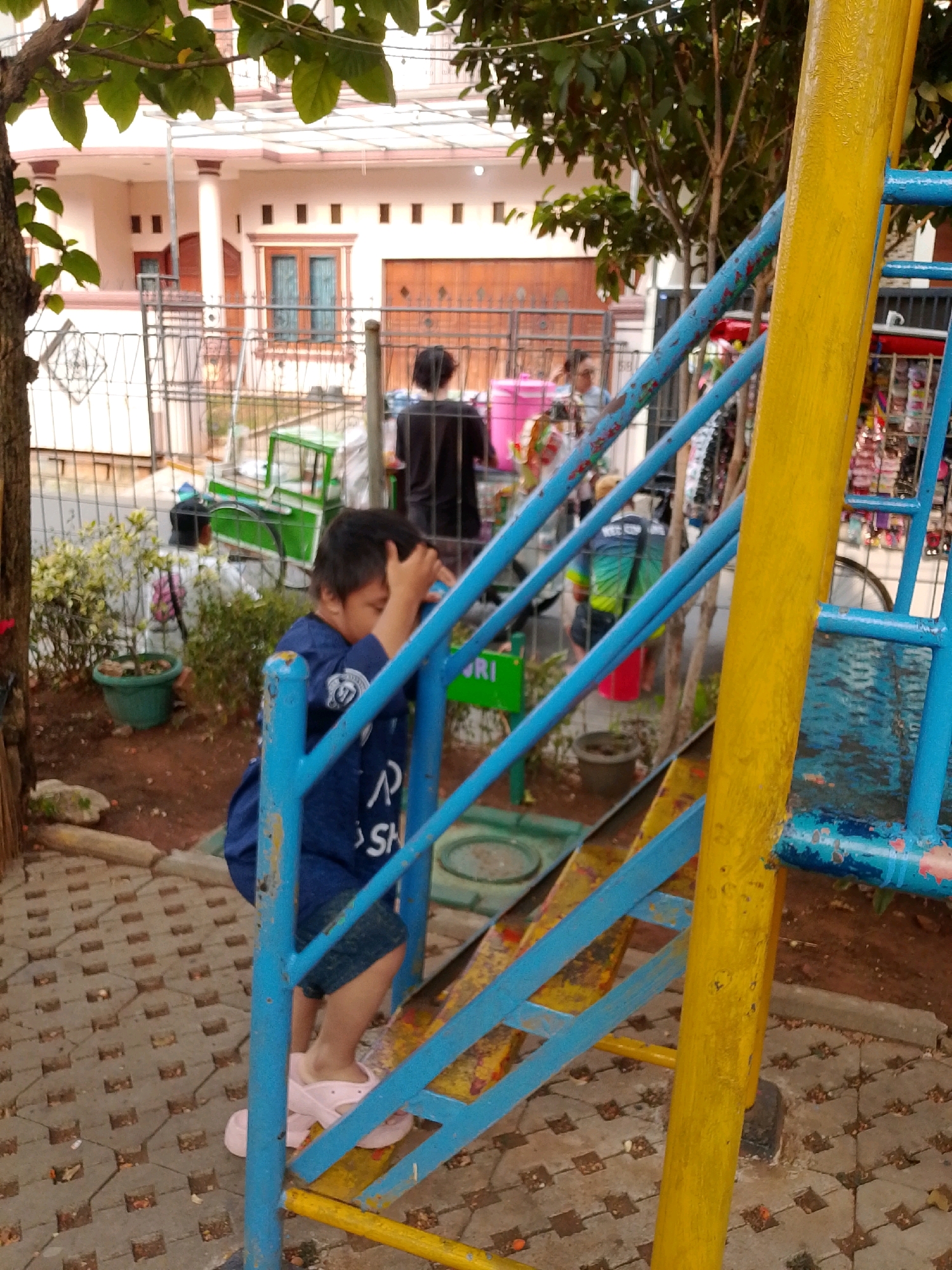 In East Jakarta, Indonesia, a young child in a blue shirt and denim shorts is playing on a playground structure. The child is facing away from the camera, with their right hand raised to their head. The playground equipment is made of blue and yellow metal. The ground is paved with grey interlocking bricks, and dirt is visible in patches. Behind the playground, a white house with a brown roof and wooden doors and windows is visible. A metal fence separates the house from the playground area. Several trees and green plants are scattered throughout the scene. Other people are visible in the background, though their faces are not clear. A green cart with a white awning is also present in the background. The overall setting appears to be an outdoor residential area with a playground.